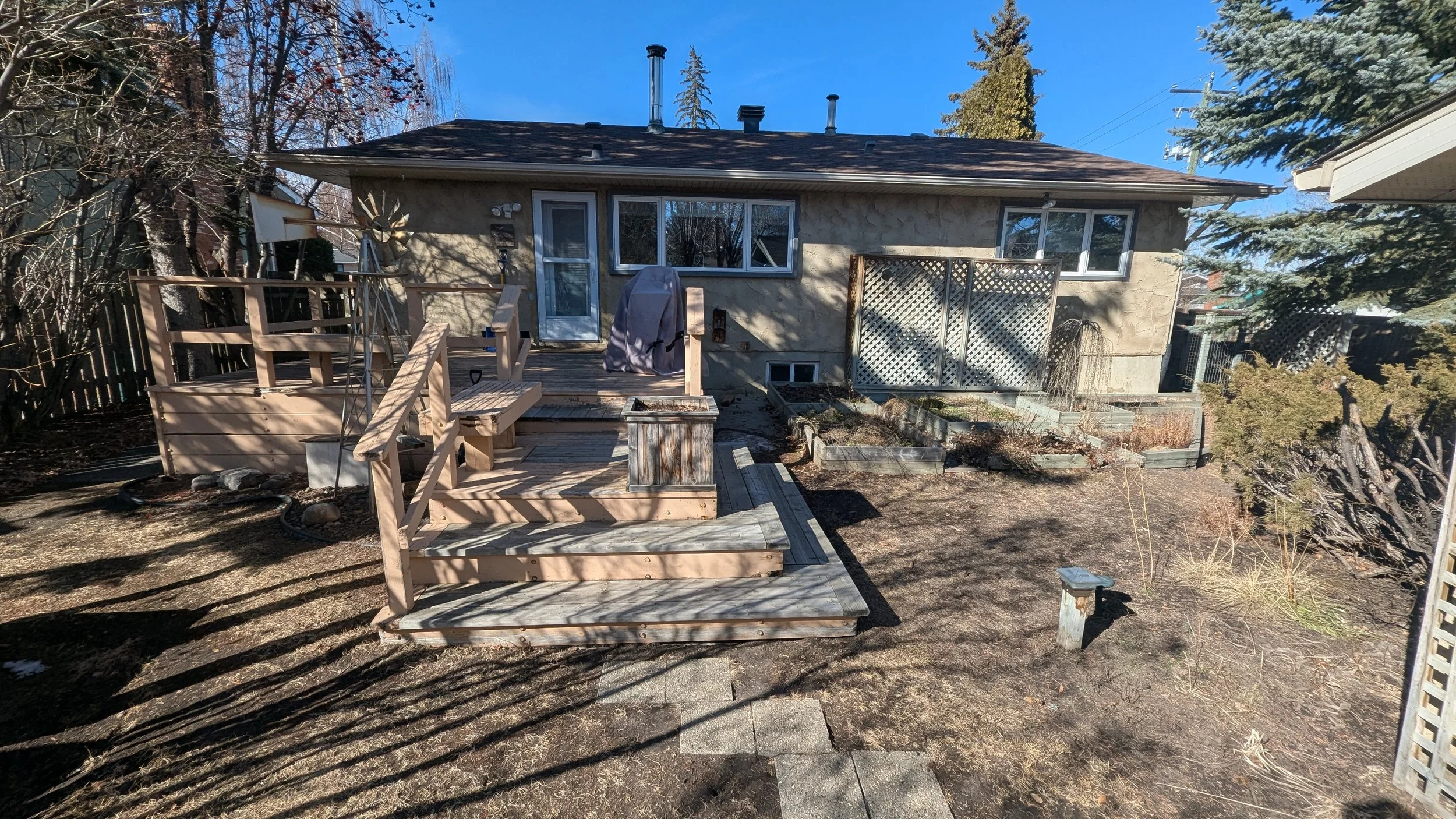 Backyard view of a beige house with a wooden deck and stairs. Uncovered soil and sparse plants are visible in the yard, with a small stone pathway leading to the deck. Tall trees and a fence are in the background, and the sky is clear and sunny.
