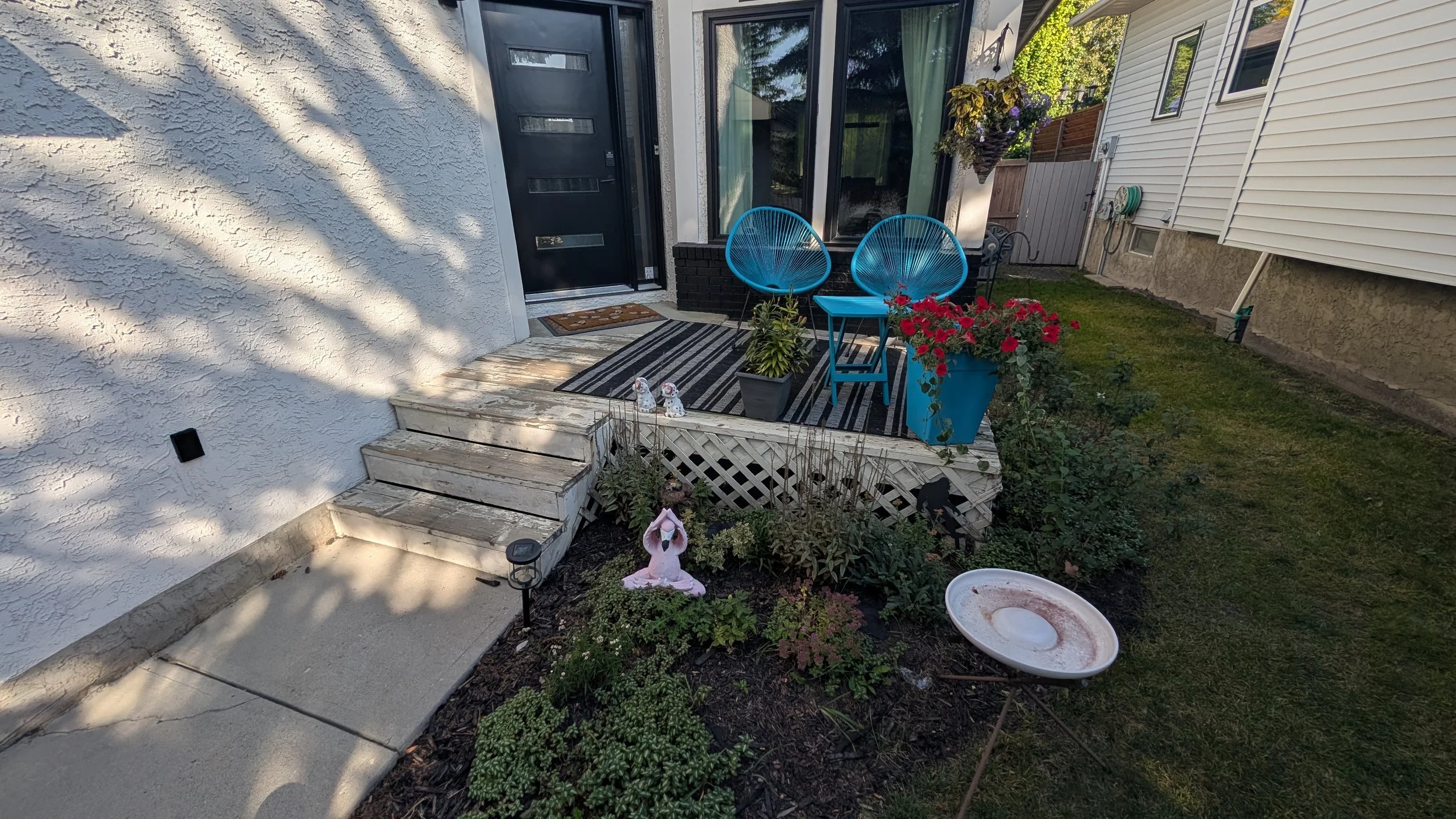 Small outdoor porch with two blue chairs, a small blue table, potted plants, a striped rug, and garden decorations in a backyard with grass and plants.
