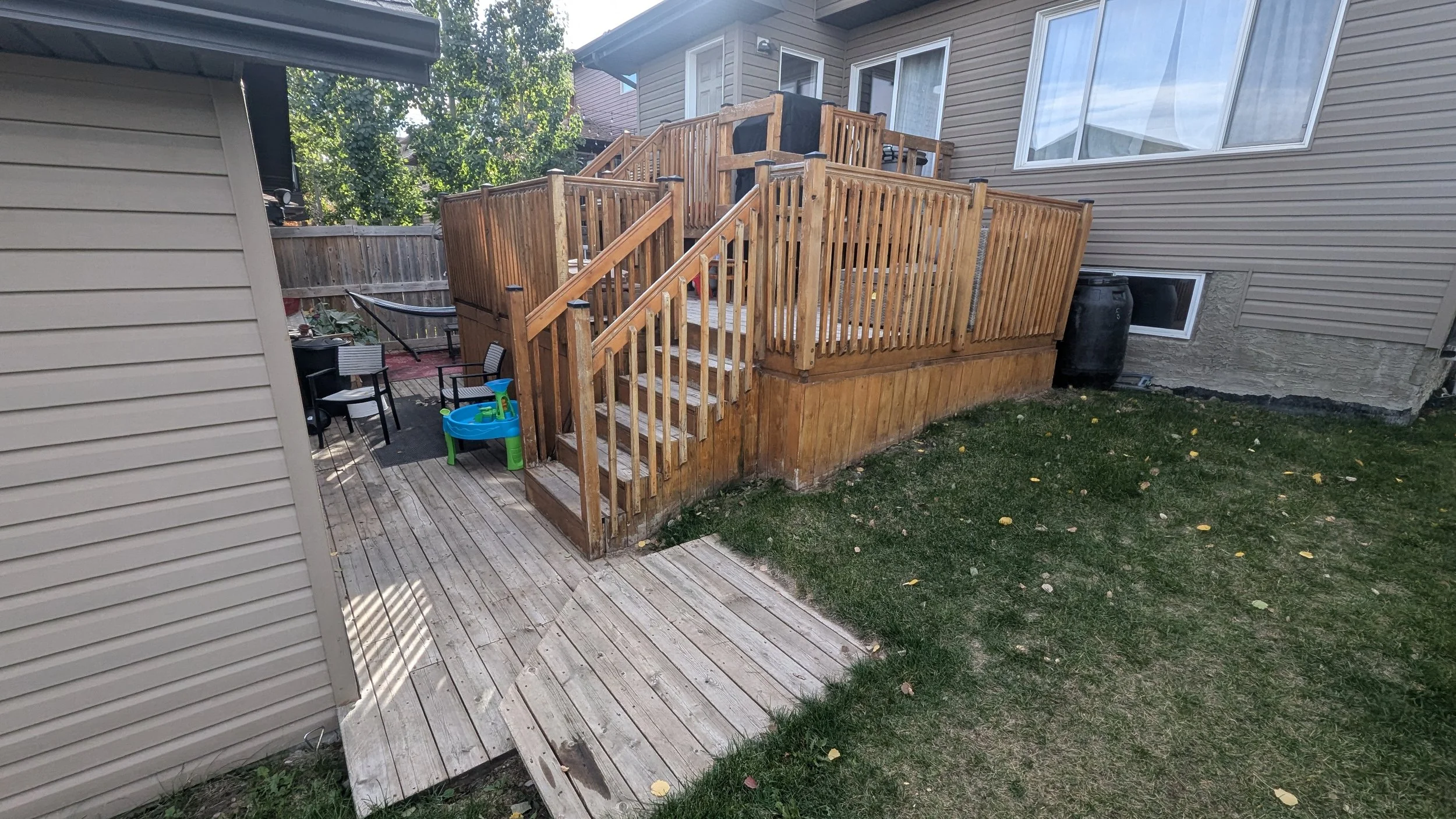 Wooden backyard deck with stairs, adjacent to a house with beige siding, small grassy area, and outdoor furniture including a blue plastic table and black chairs.