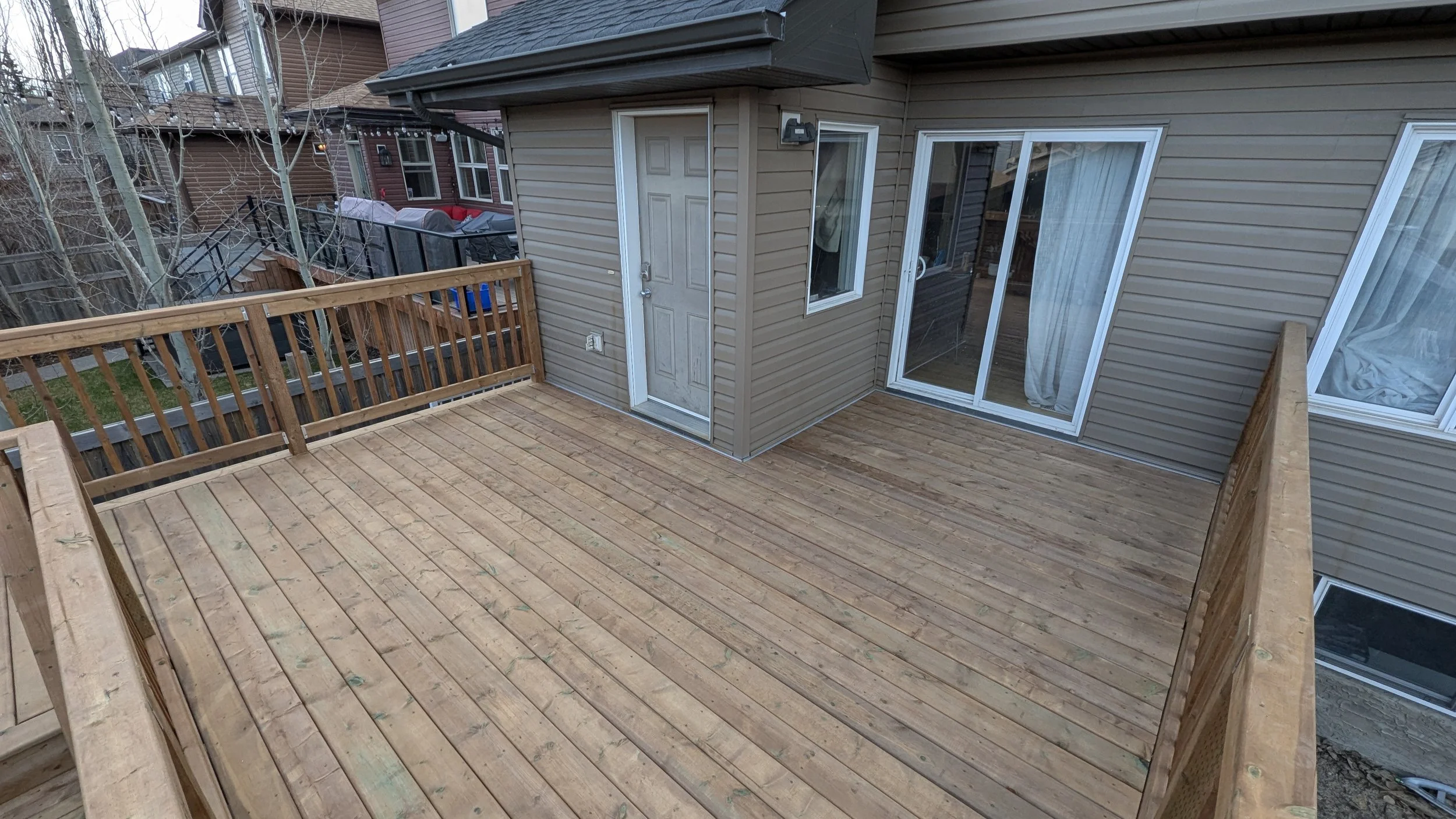 Newly constructed wooden deck attached to a house with beige siding, featuring a door, sliding glass door, and two windows, with a neighboring deck and backyard visible in the background.