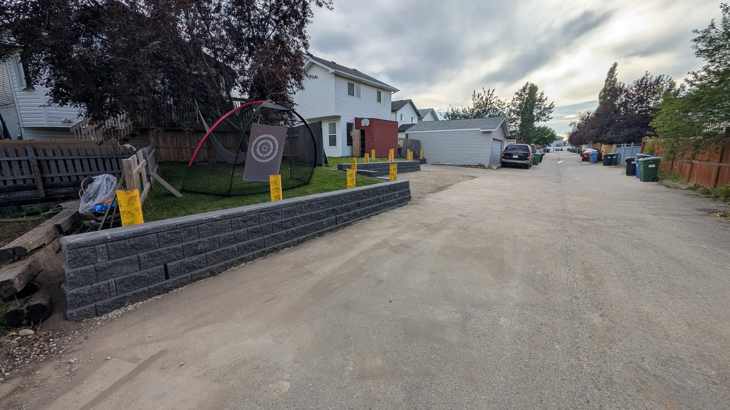 A residential street with a partially built stone retaining wall and yellow construction warning signs along it. Houses with fenced yards and several parked cars are visible in the background, under cloudy skies.