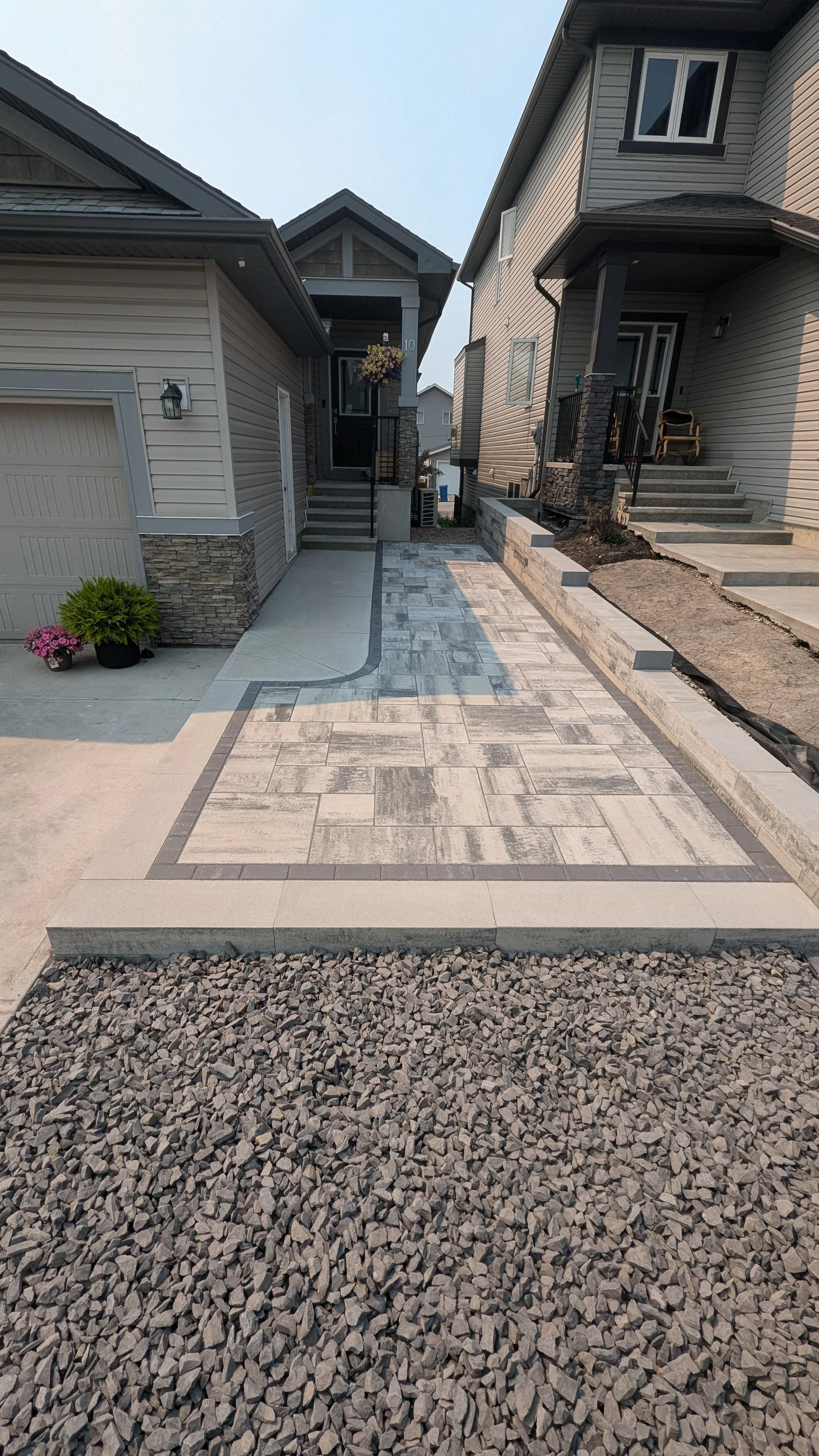 A newly paved driveway with patterned concrete and gravel on each side, leading to the front entrances of two houses in a suburban neighborhood.