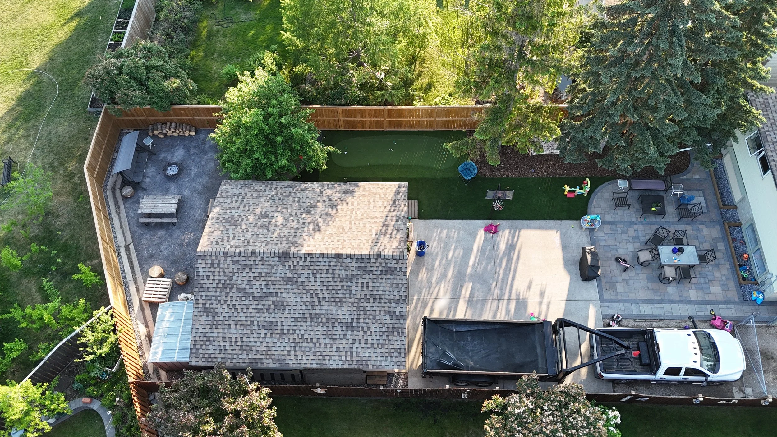 An aerial view of a backyard with various sections including a stone patio with outdoor seating, a grassy area, a small deck with children’s toys, and a parking space with a white truck. Tall trees and a wooden fence surround the yard.