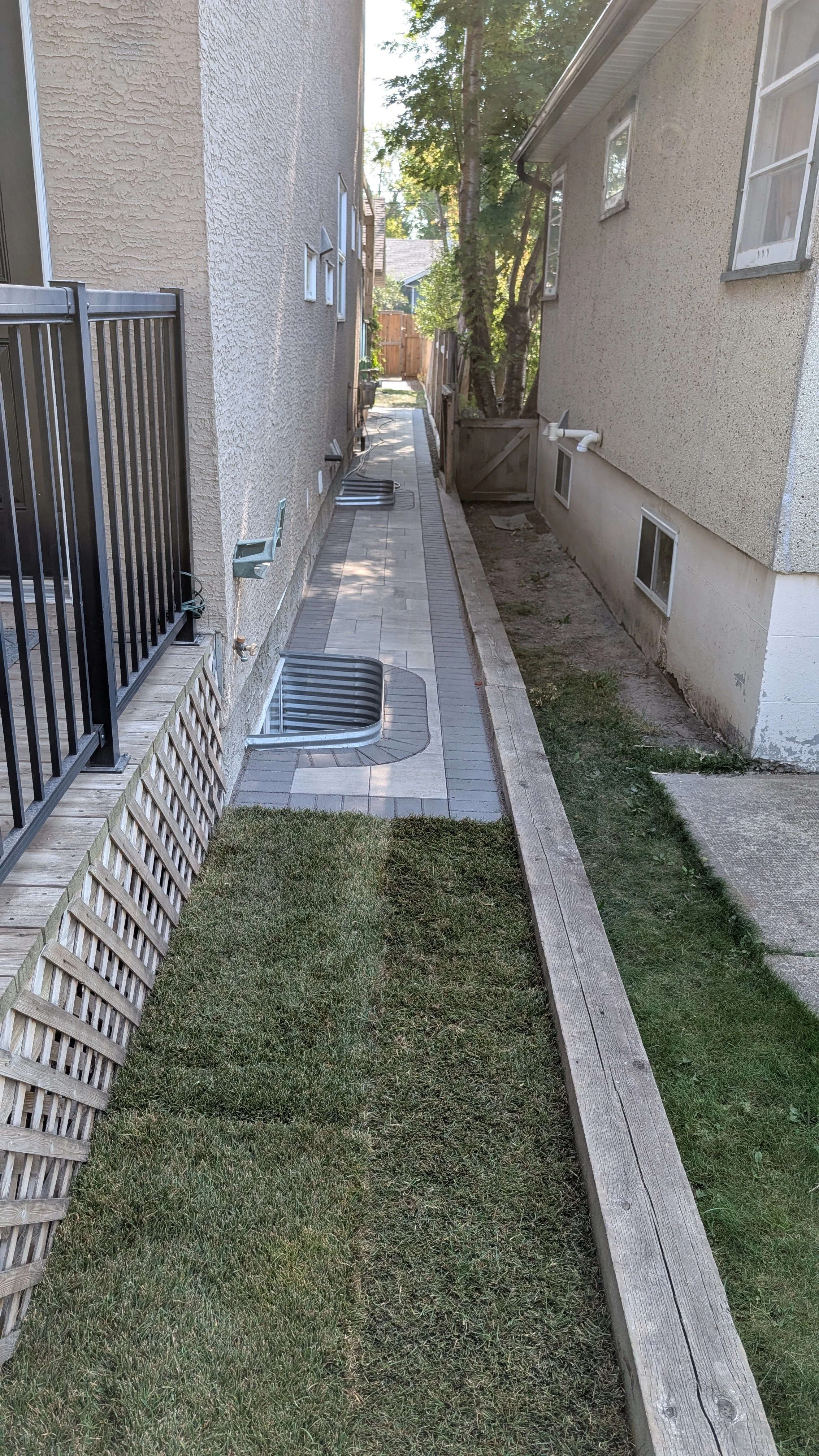 Side yard with a small grassy area, wooden border, concrete walkway, and exterior walls of two houses with windows.