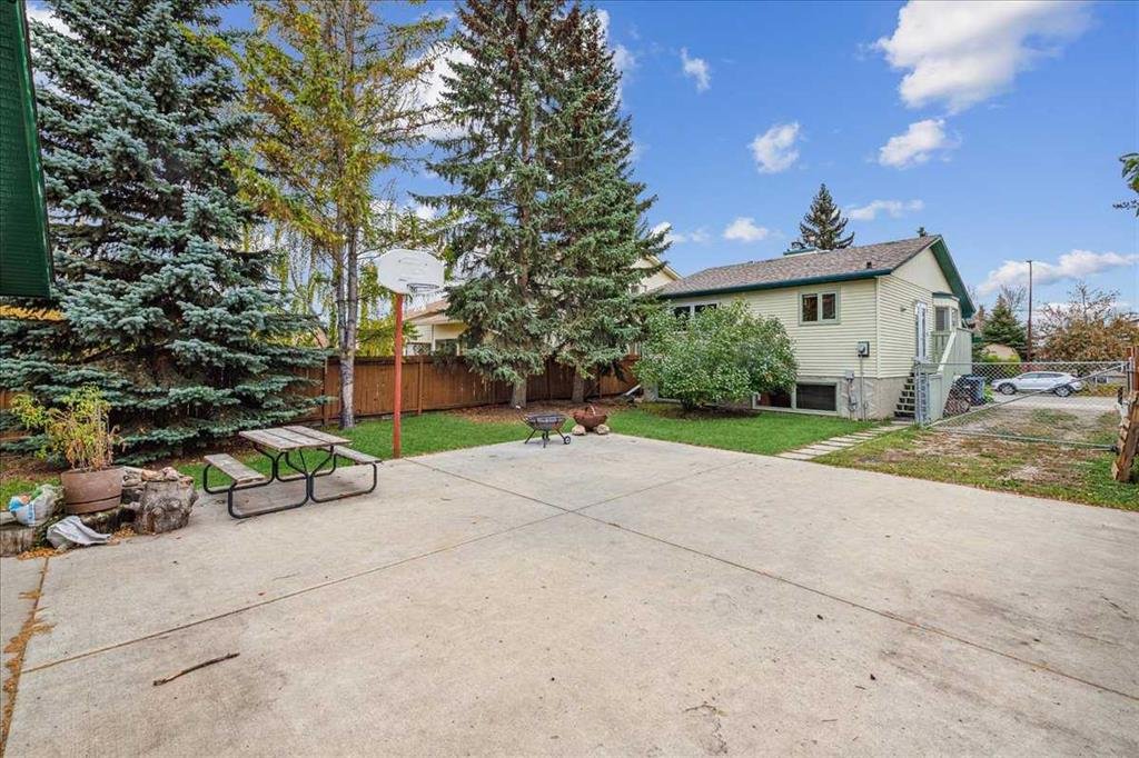 A backyard with a concrete patio, picnic table, a basketball hoop, small grassy area, trees, and a house with stairs leading to the backyard.