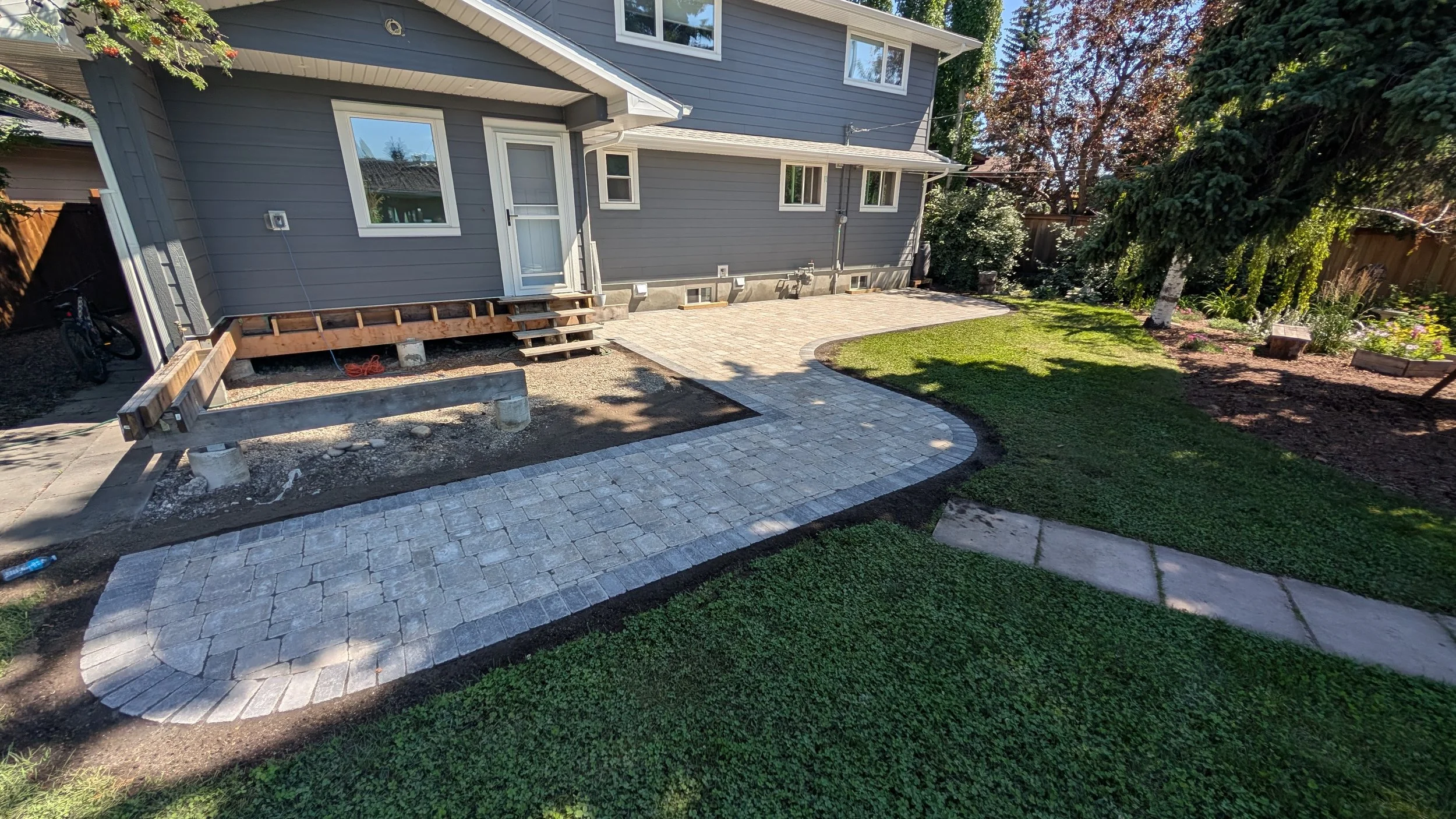 Backyard with a newly paved stone pathway leading to a gray house, with a grassy lawn and flower beds on the right, and trees casting shadows.