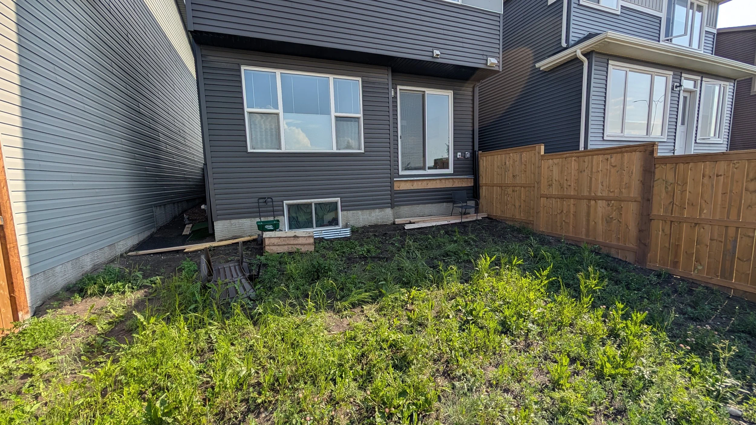 Backyard of a townhouse with fenced backyard, blue and gray siding, a small garden area with weeds, a wooden fence, and construction materials near the house.
