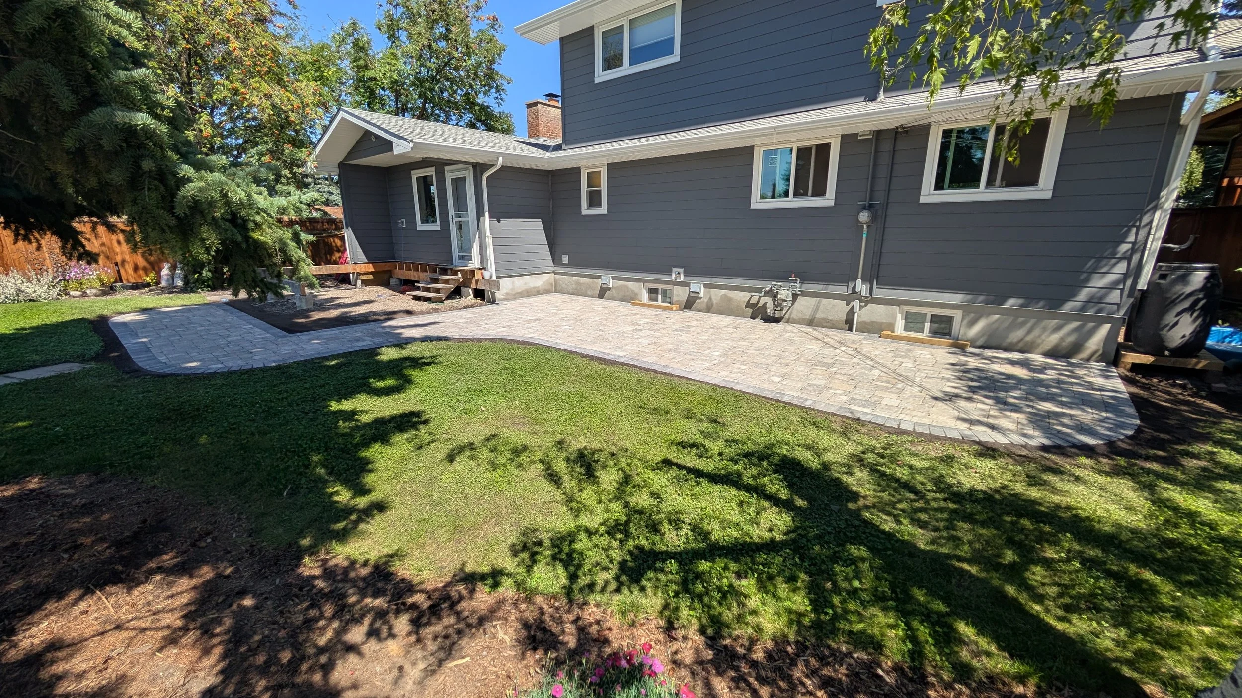 Backyard with newly installed stone patio, green grass, trees, and a gray house with white framing.