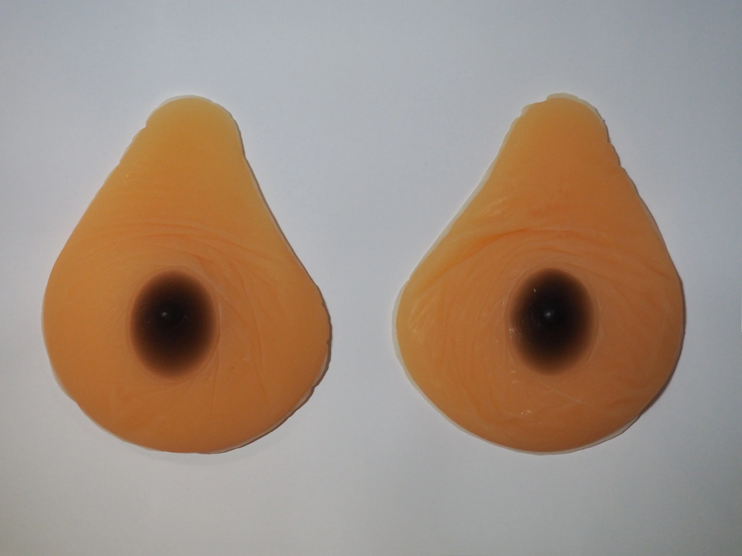 A pair of prosthetic breasts. They are pear-shaped, and have pale skin and dark brown nipples and aureolas. There are some creases in the silicone around the nipples.