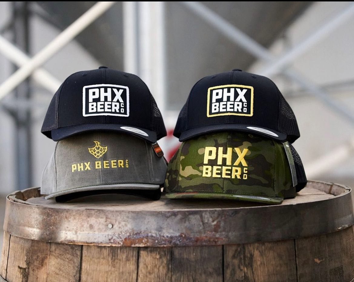 Four snapback hats with embroidered logos saying PHX BEER CO sitting on a beer keg