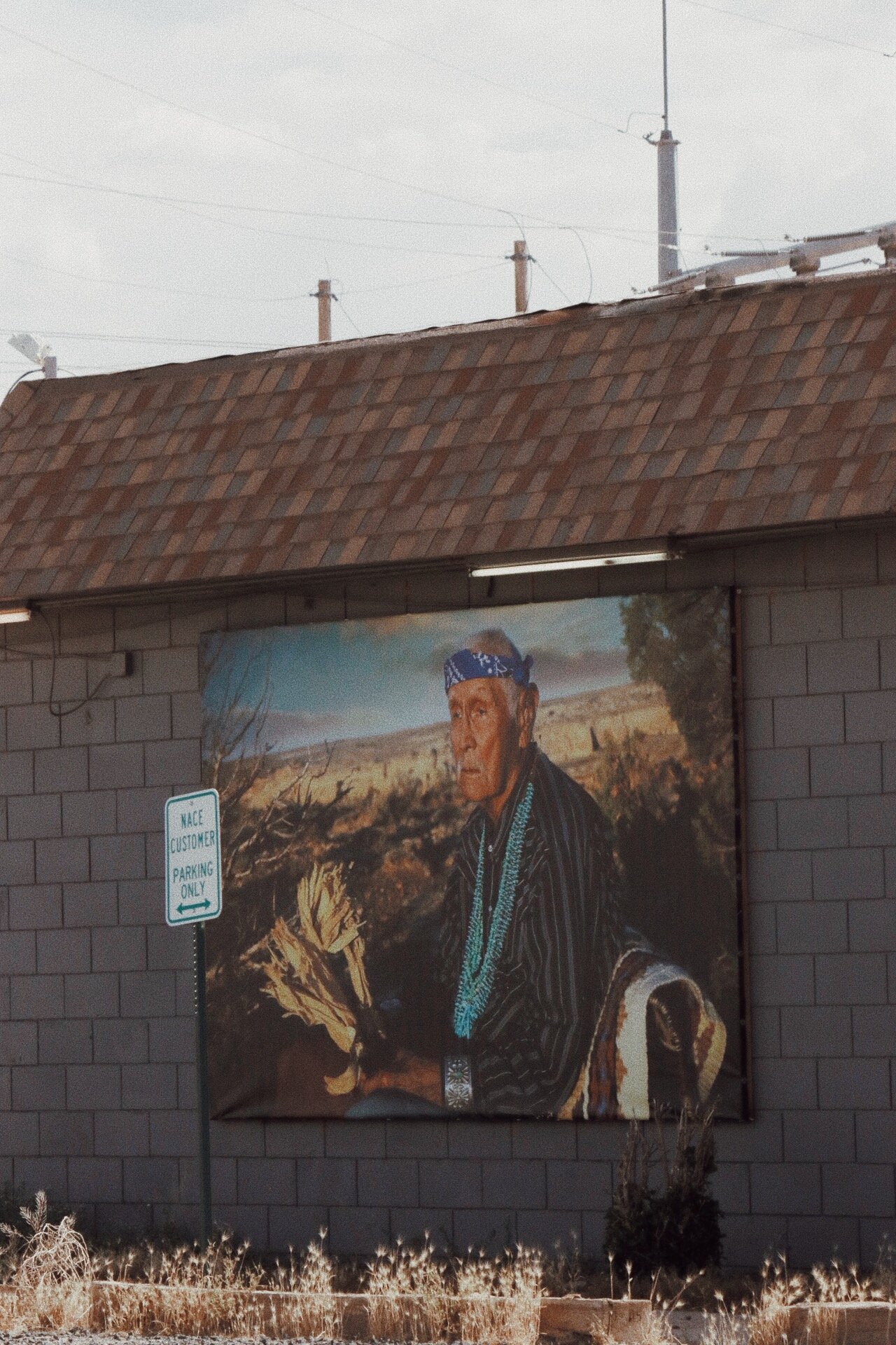 A large mural of an elderly Indigenous man wearing a blue bandana and turquoise necklaces, dressed in traditional clothing, outdoors in a desert landscape, on the side of a building.