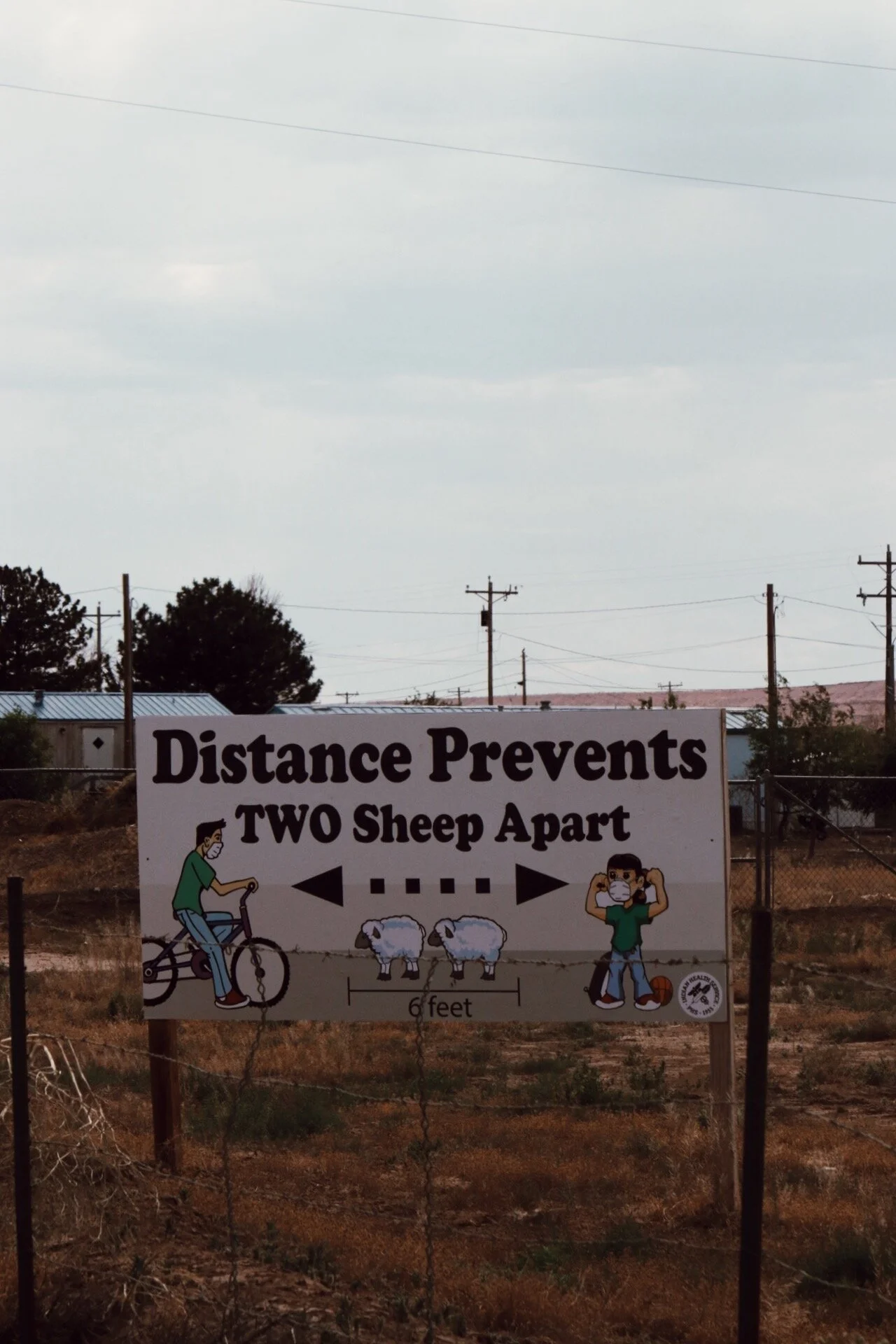 A sign indicating social distancing with the message "Distance Prevents Two Sheep Apart" depicting two sheep separated by six feet, and illustrations of a person riding a bike and a person adjusting a face mask, both maintaining distance.