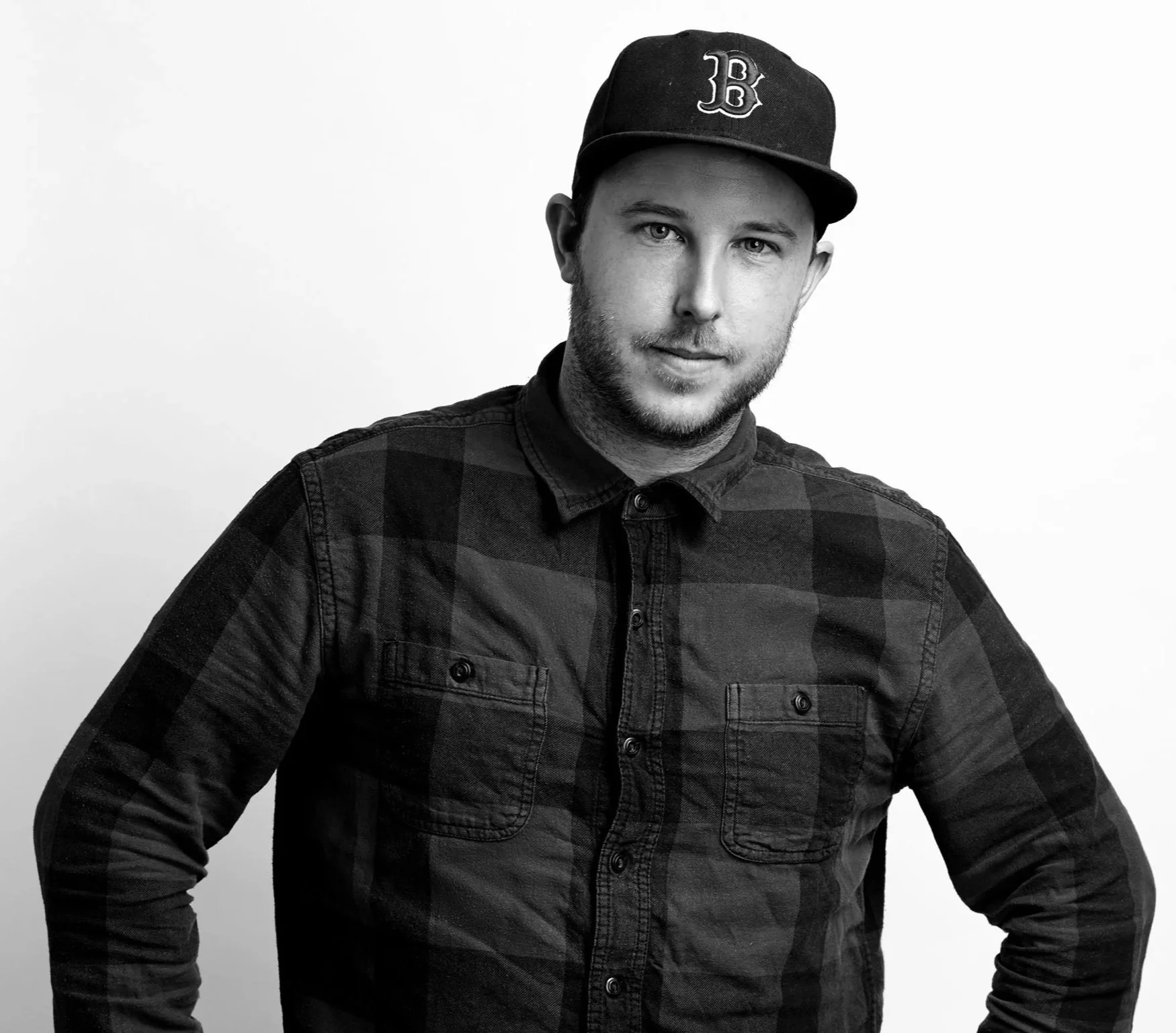 Black and white portrait of a young man wearing a baseball cap with a 'B' logo, a dark plaid button-up shirt, standing against a plain background.