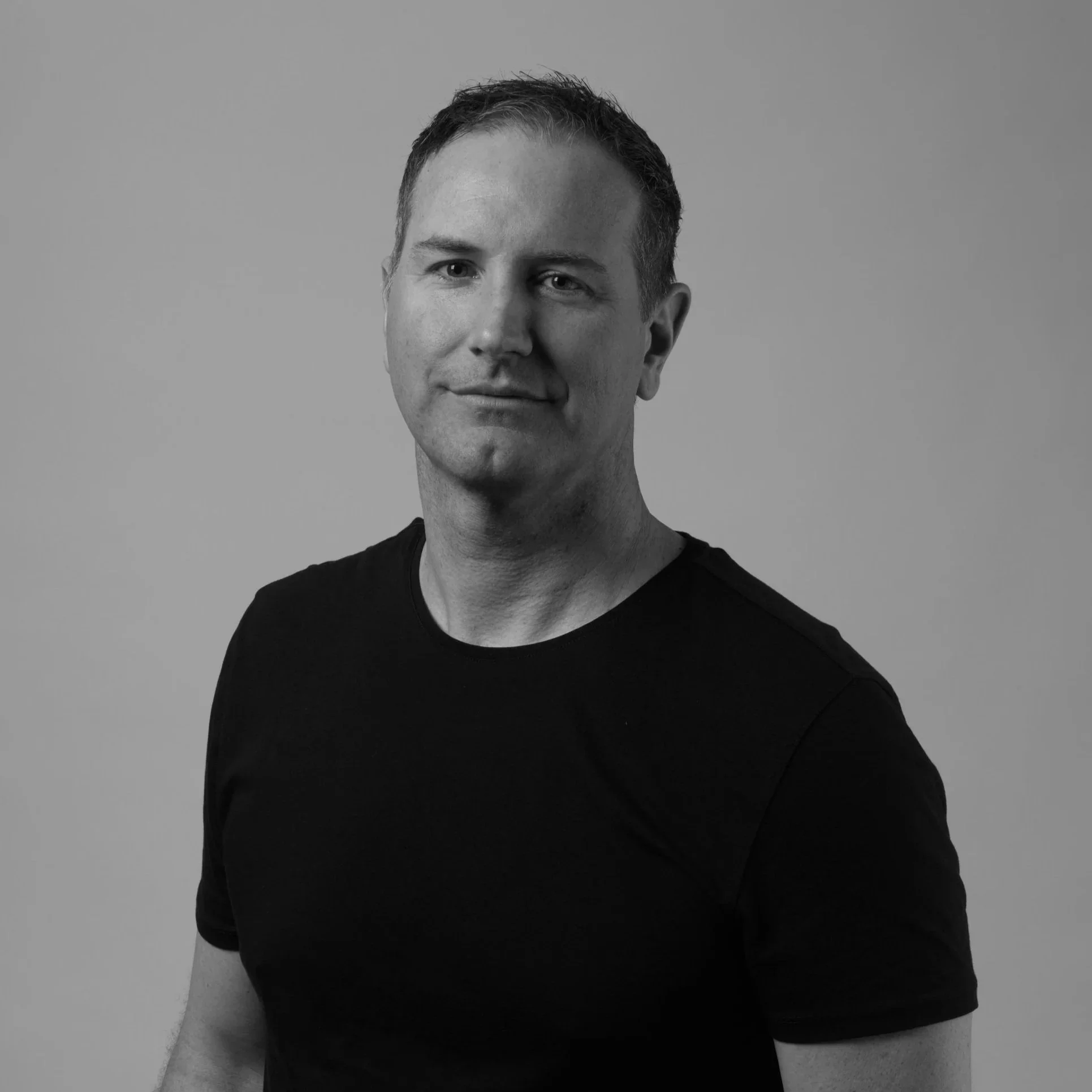 Black and white portrait of a man wearing a black T-shirt, looking at the camera with a slight smile, on a plain background.