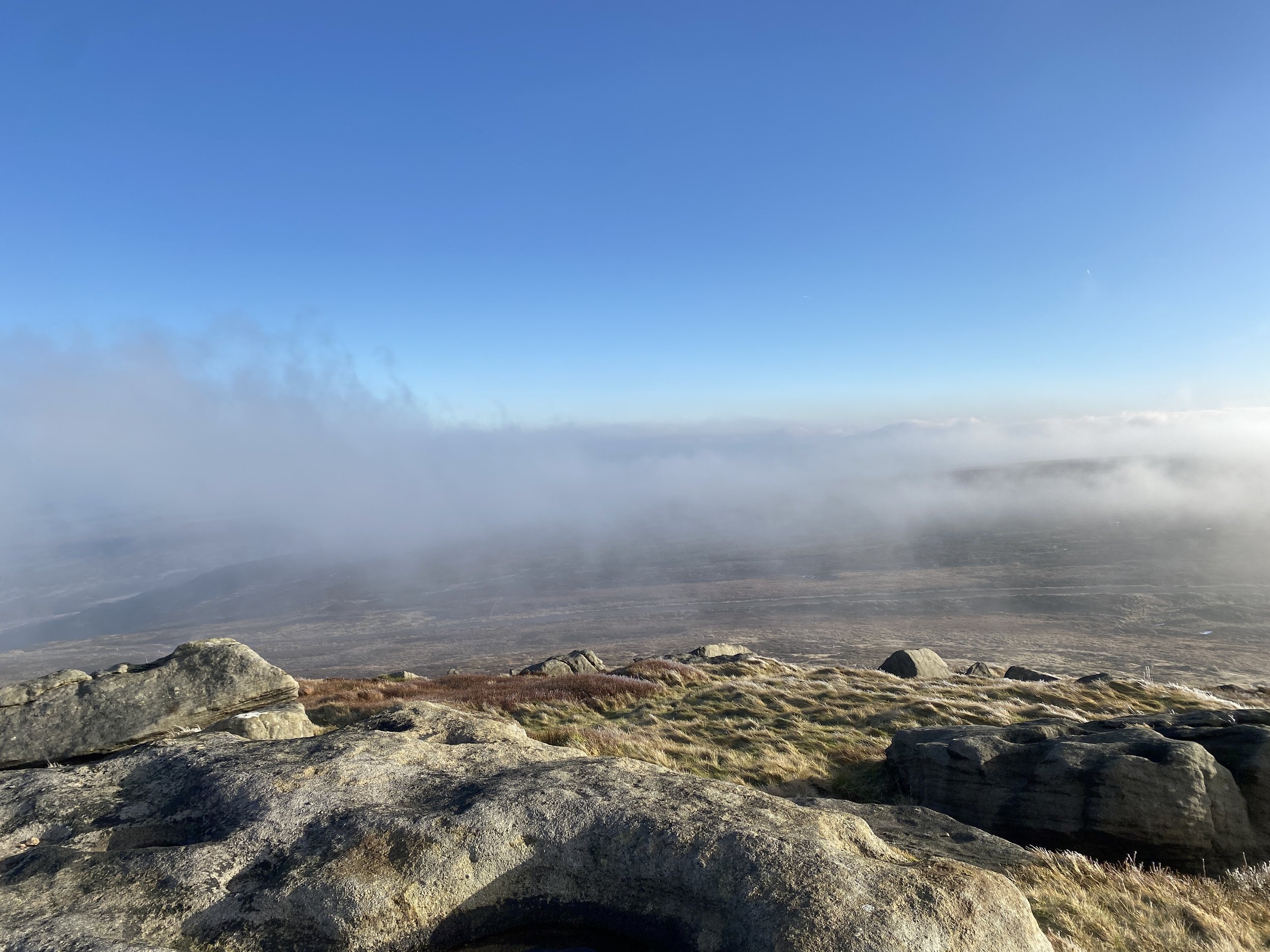 View from West Nab Summit, rocks in the foreground, looking out over moorland. Blue skies above and cloud blowing across the moorland.