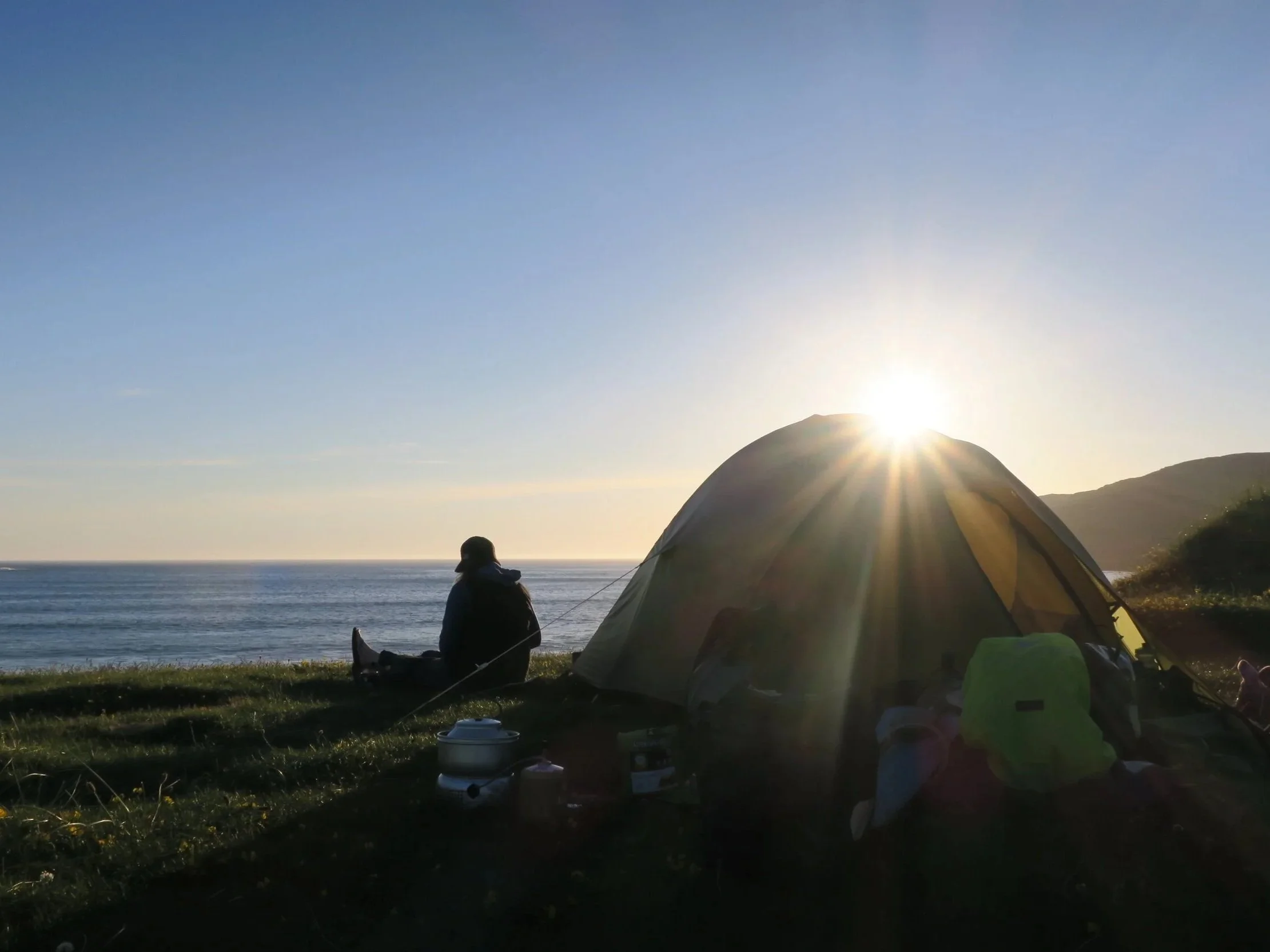 Person sitting outside a wild camp at sunset, looking out over the sea