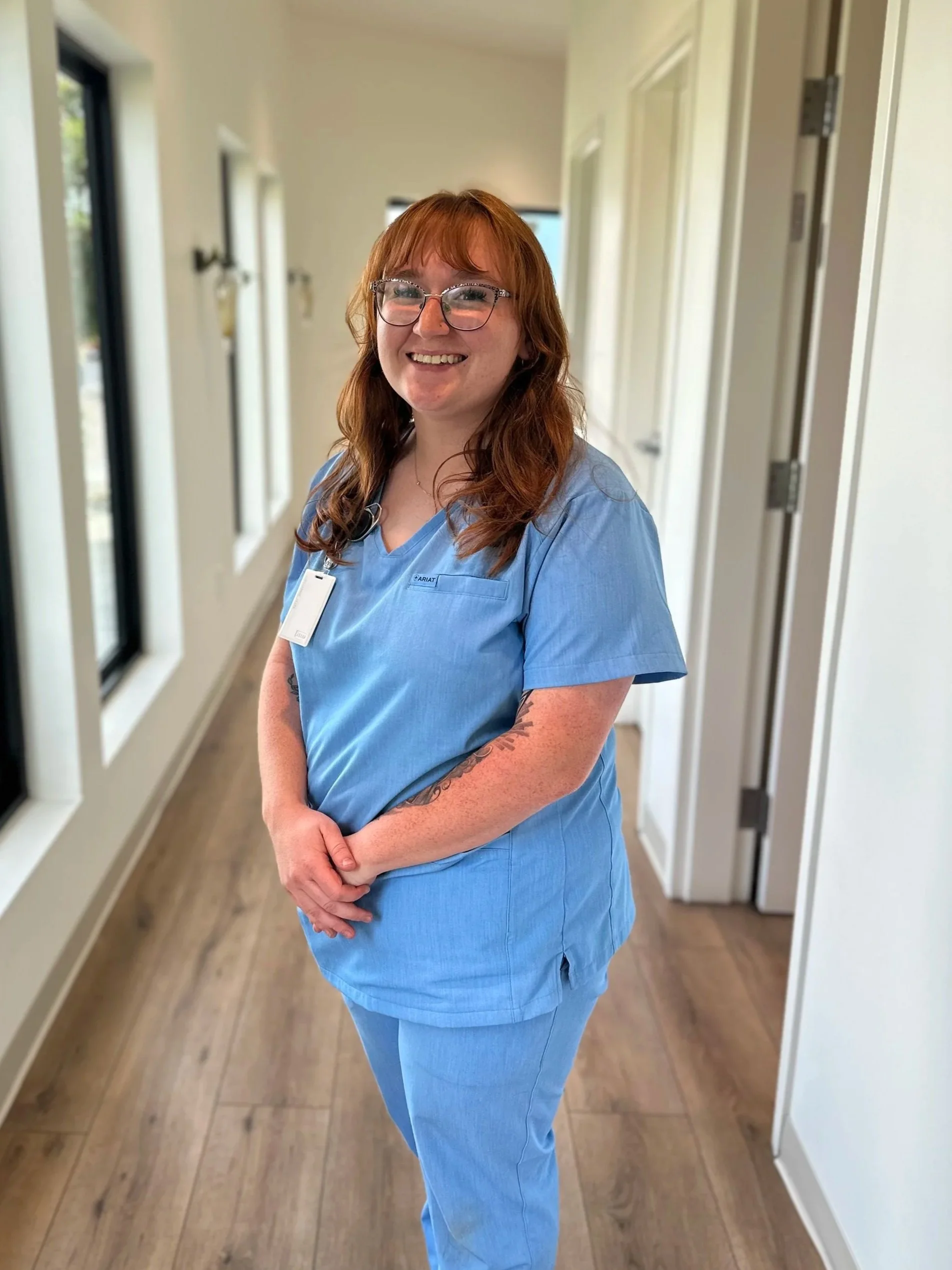 Person wearing teal medical scrubs in a bright hallway, smiling.