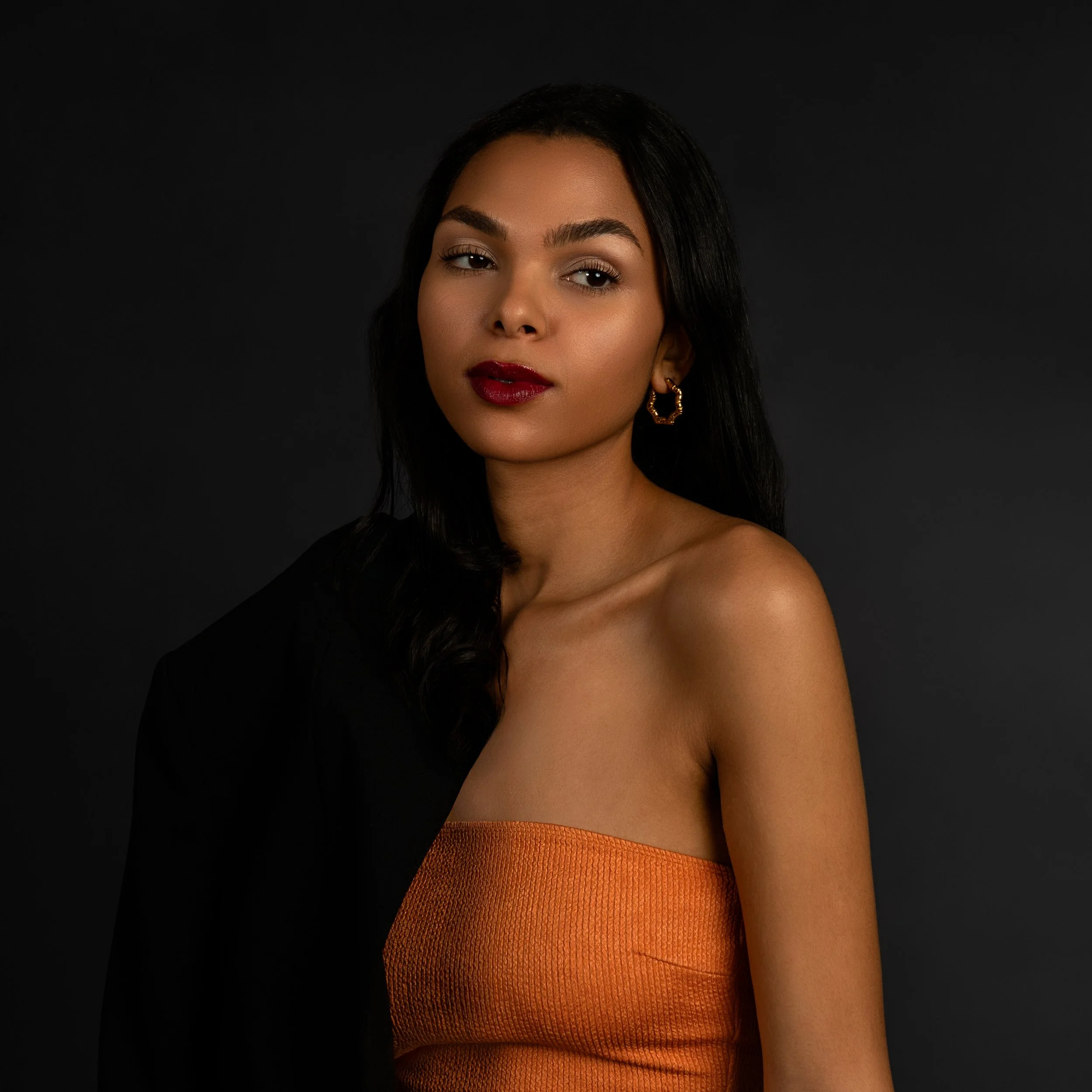 Woman with dark hair in an orange strapless top, wearing hoop earrings, and a dark jacket draped over one shoulder, against a dark background.
