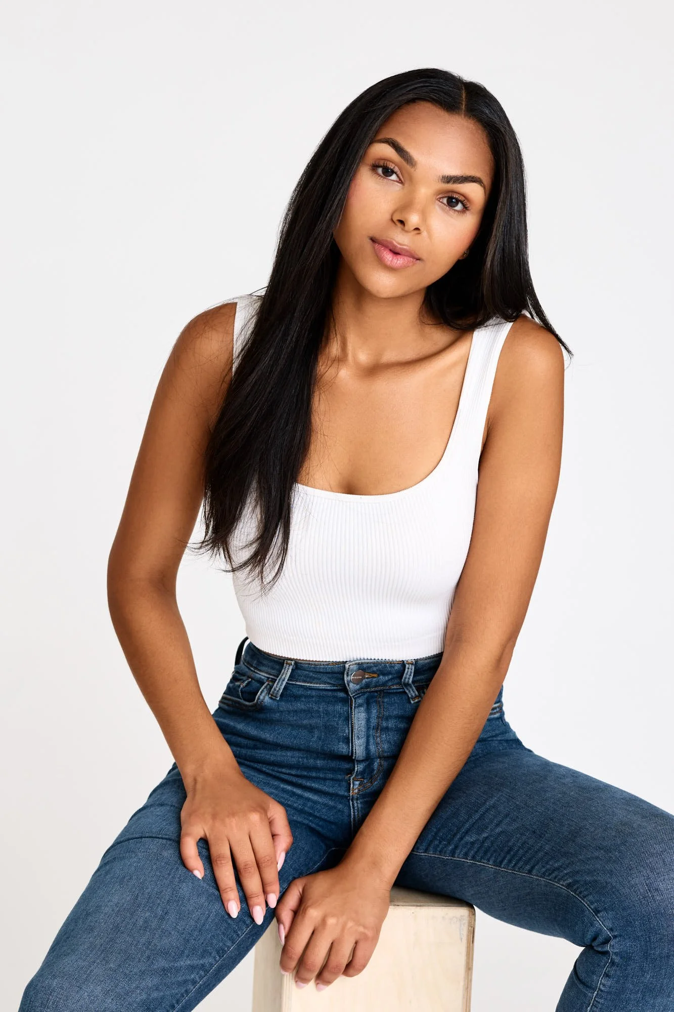 Woman with long hair in a white tank top and jeans sitting on a stool.