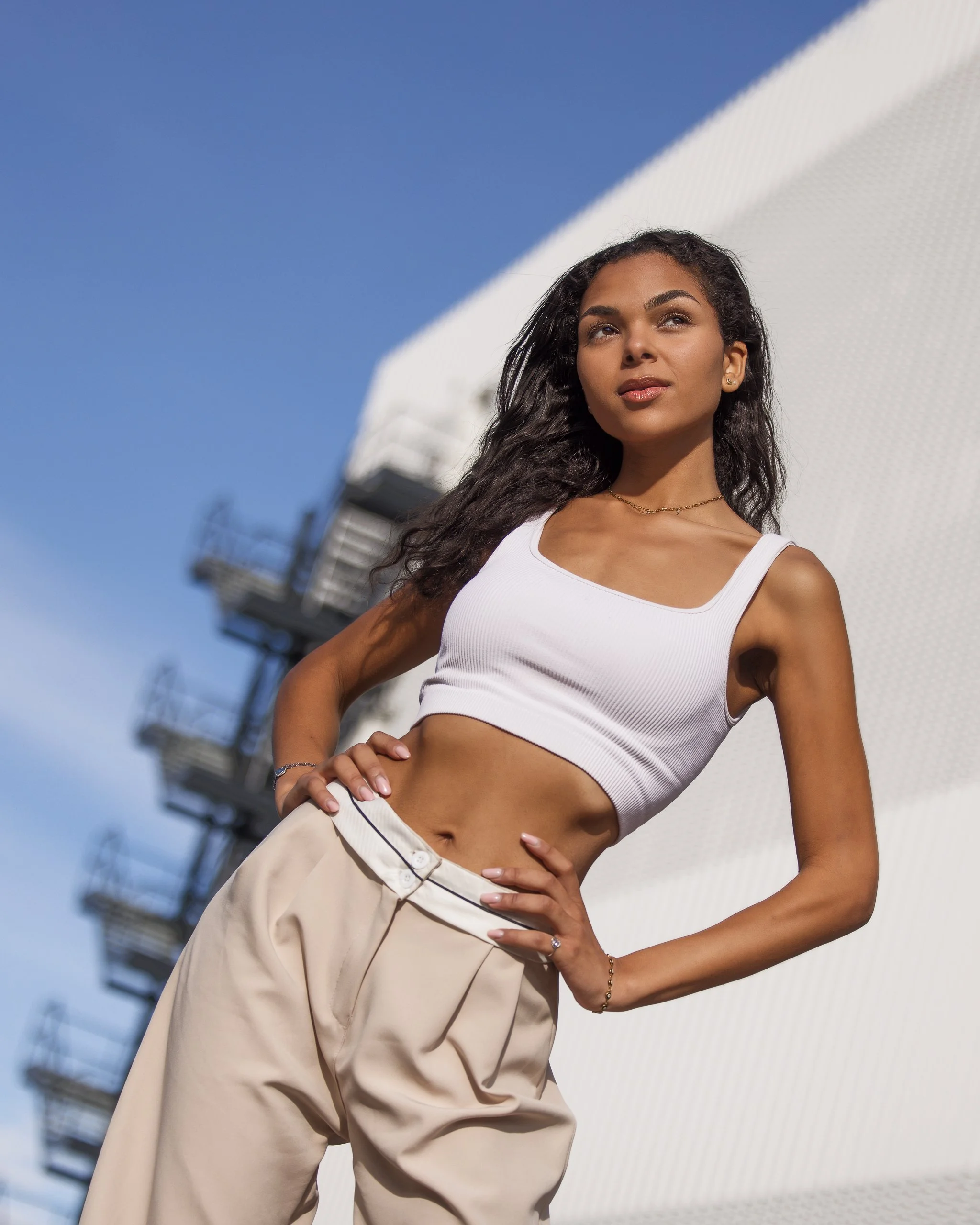 Woman in a white crop top and beige pants standing outdoors with a modern building in the background.