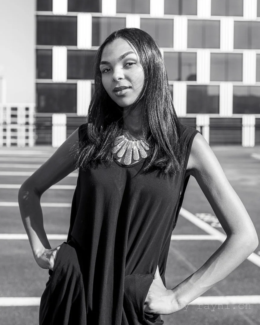 Black and white photo of a woman with long hair, wearing a necklace and a sleeveless top, standing outdoors with hands on hips, in front of a building with a geometric pattern.