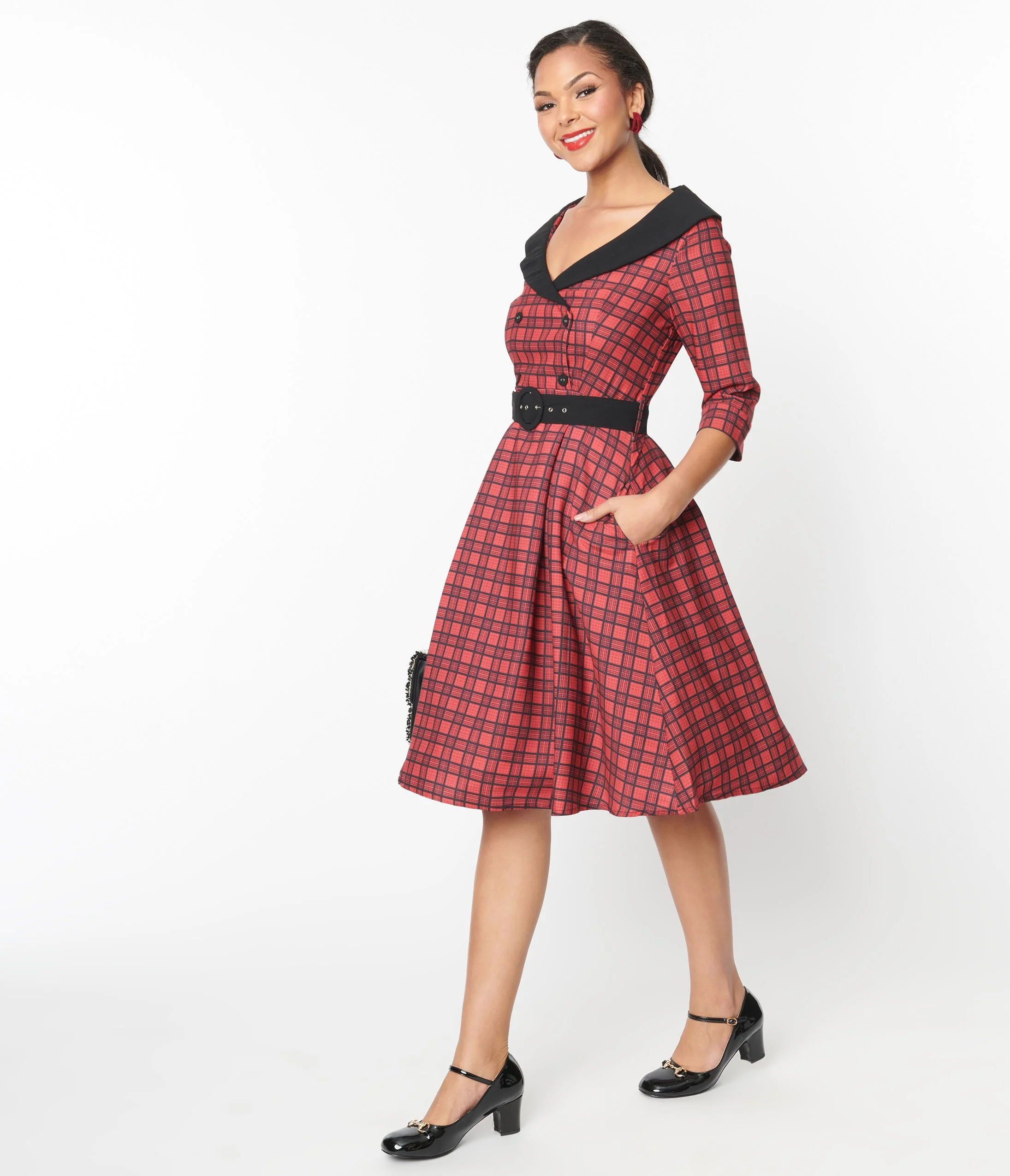 Woman in a red checkered dress with a black collar and belt, wearing black heels, standing against a white background.