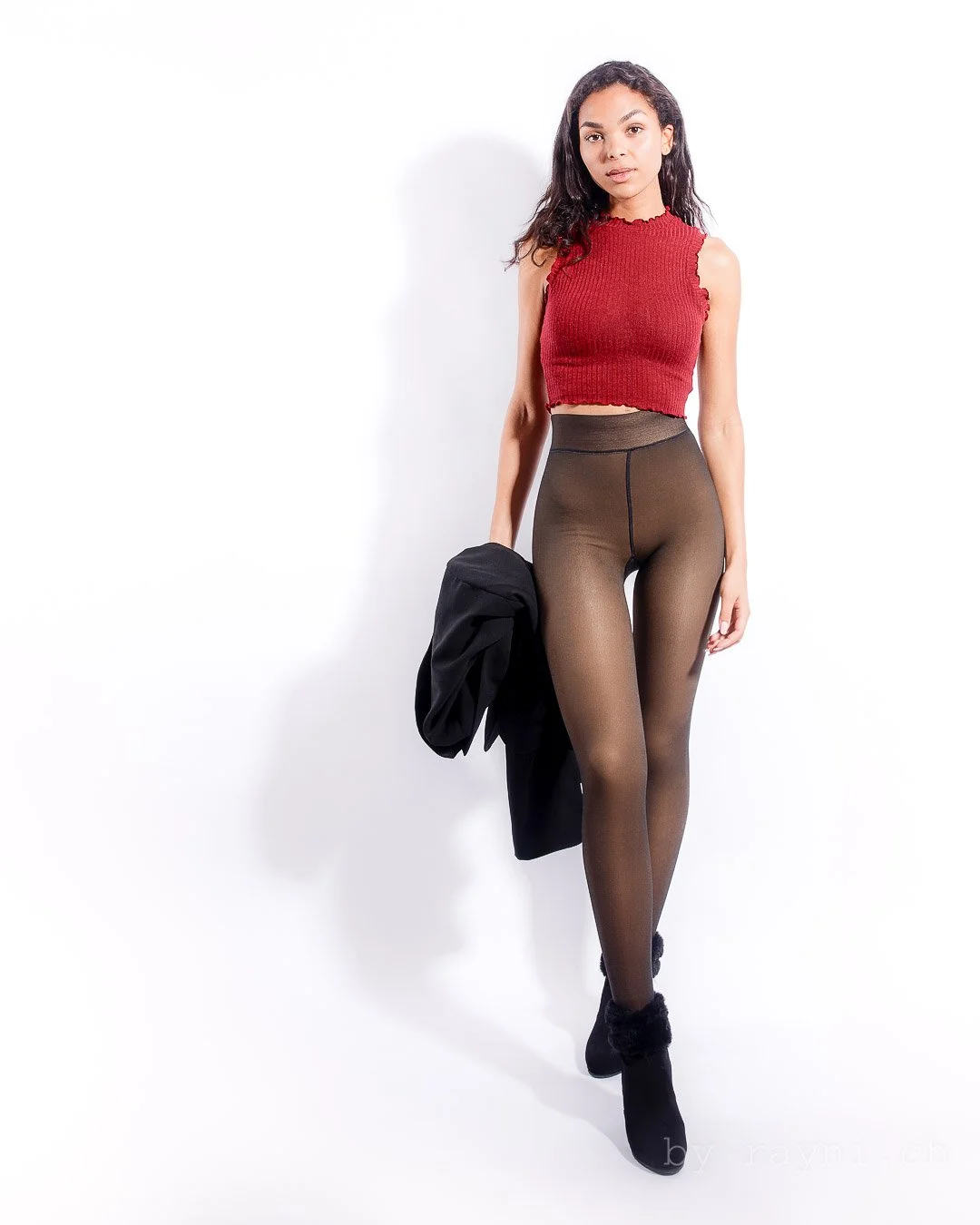 Woman in a red sleeveless crop top, sheer leggings, and black boots, holding a jacket.