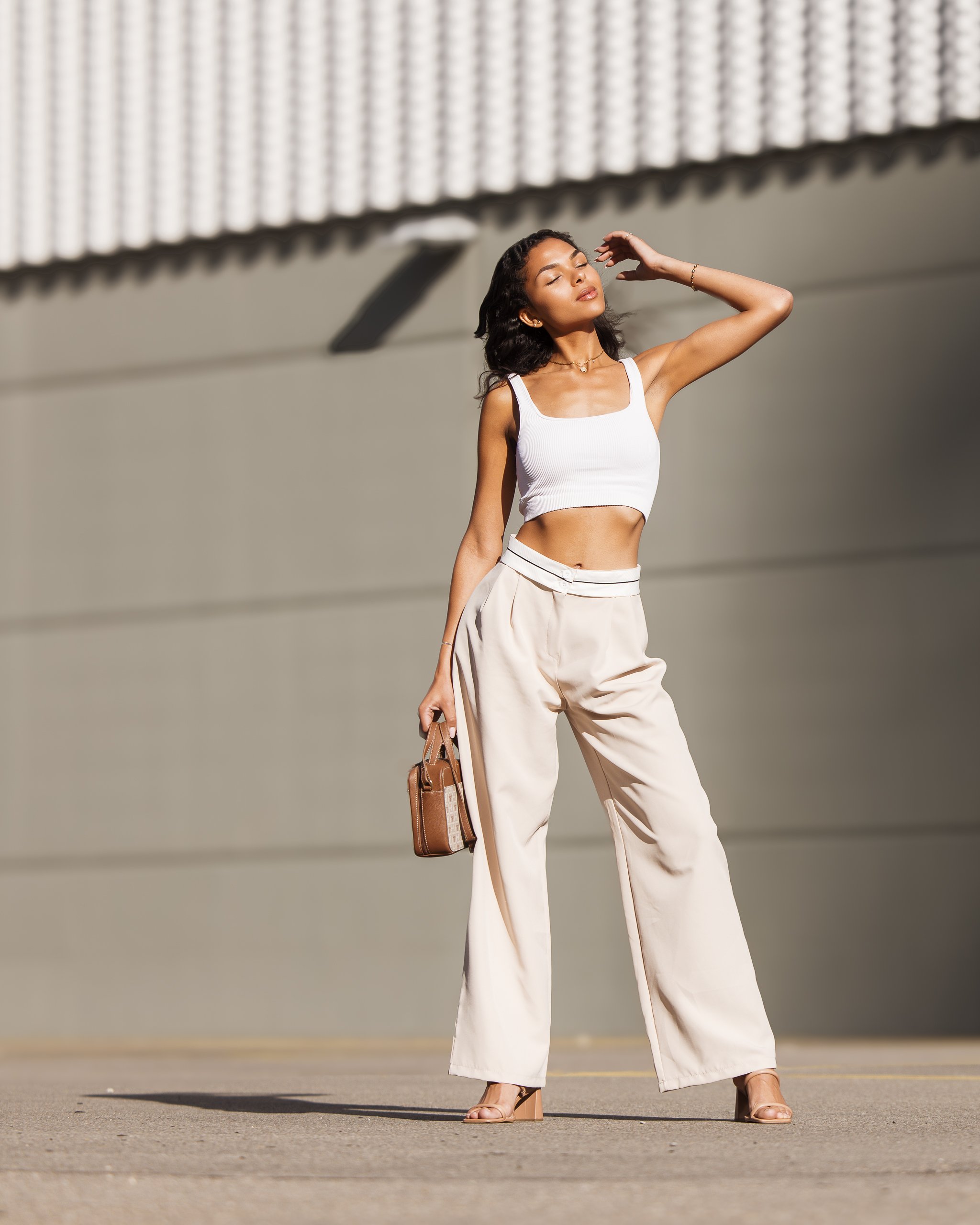 A woman standing outdoors in front of a textured wall, wearing a white crop top, beige wide-leg pants, holding a brown handbag, and posing with her hand raised to her head.
