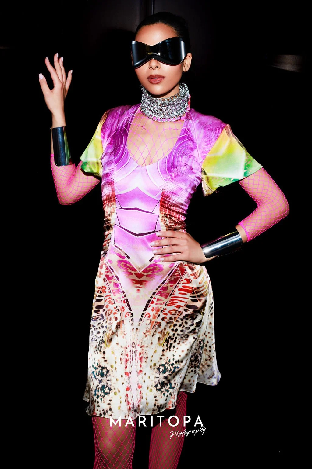 Person wearing a colorful, futuristic dress with a metallic choker, large reflective sunglasses, and silver wrist cuffs, against a dark background.