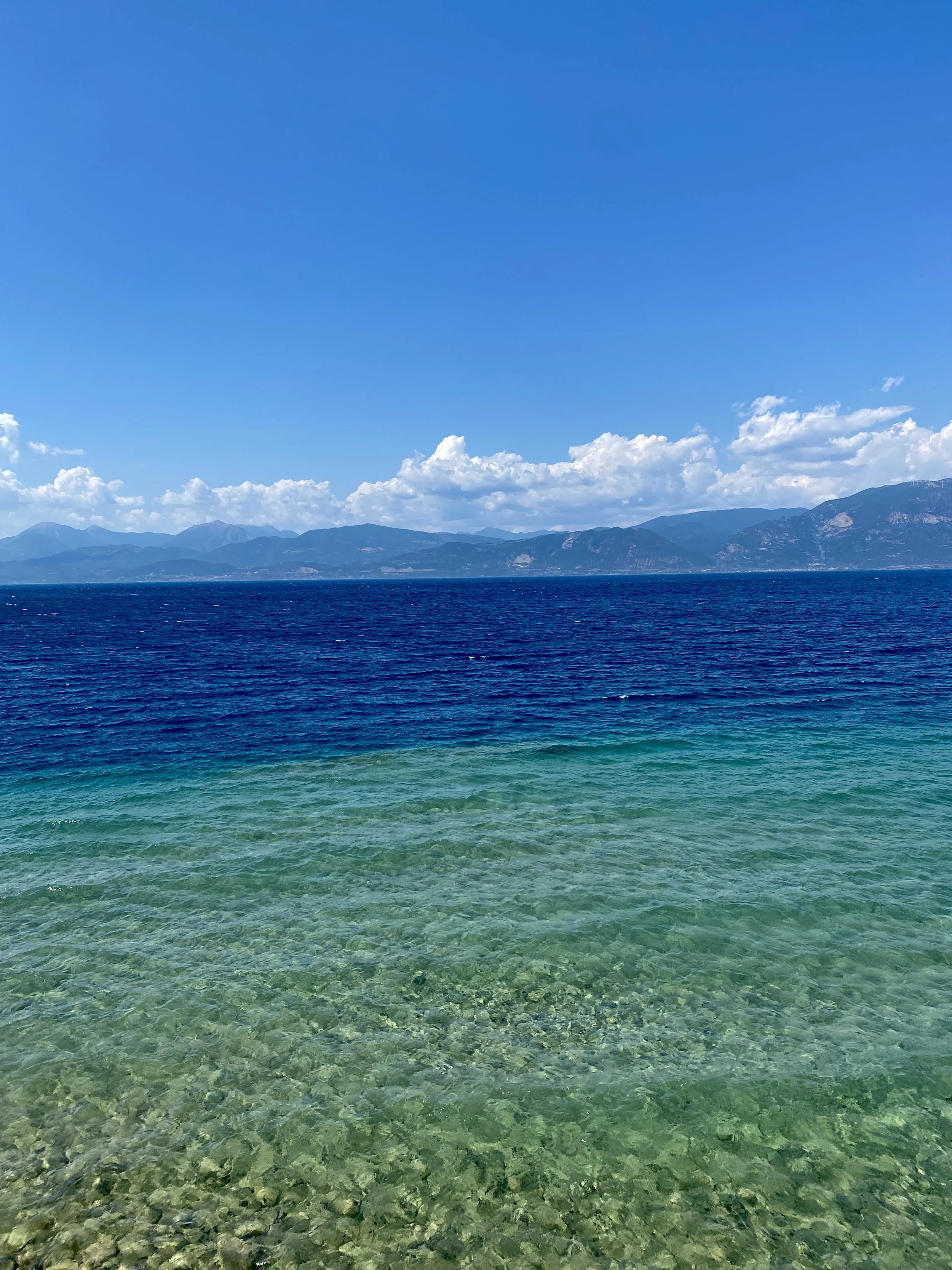 Clear ocean water with pebbles in the foreground under a blue sky with distant mountains and clouds.