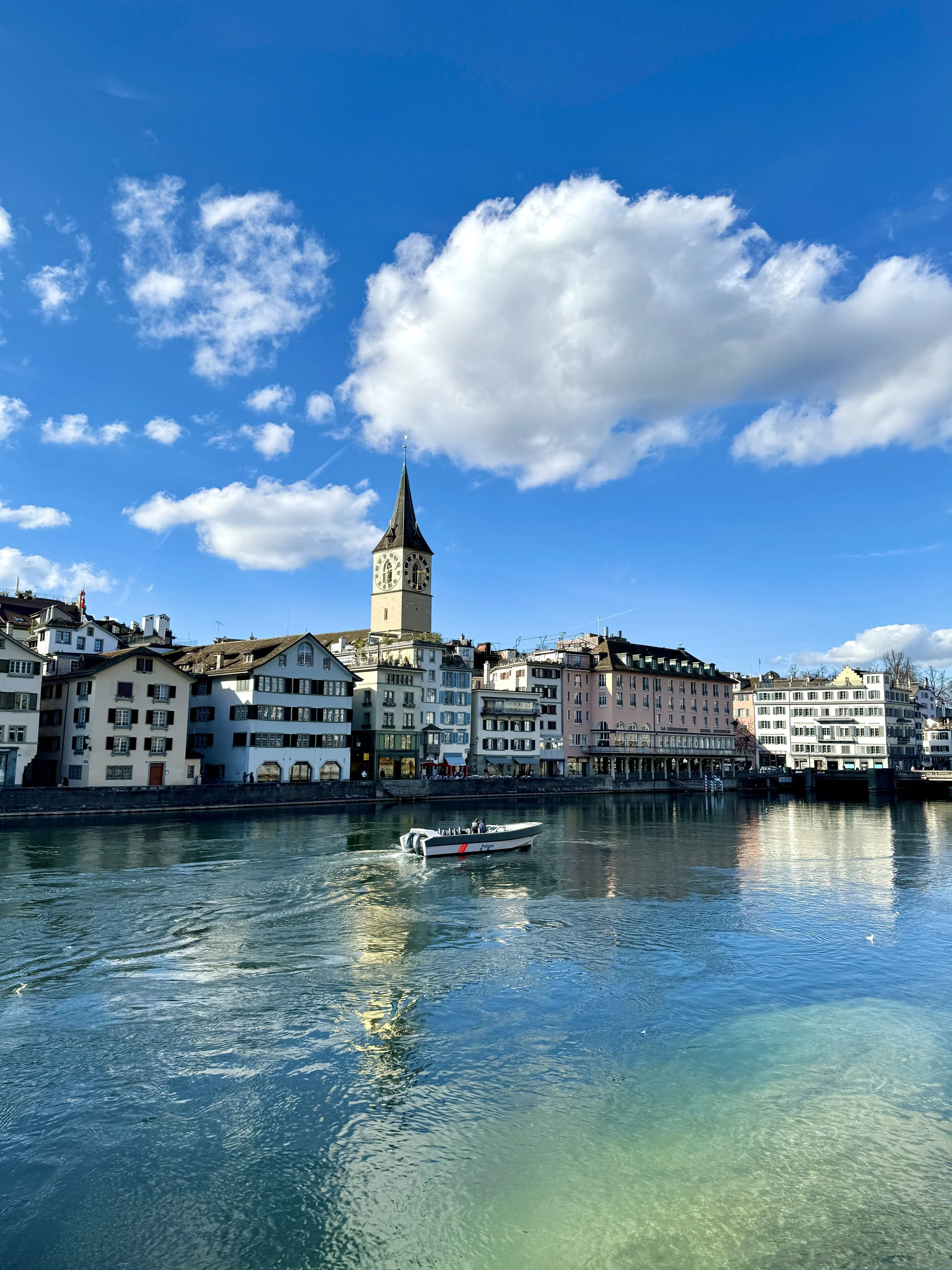 View of a river with a boat, historic buildings along the shore, and a church tower in the background against a cloudy blue sky.