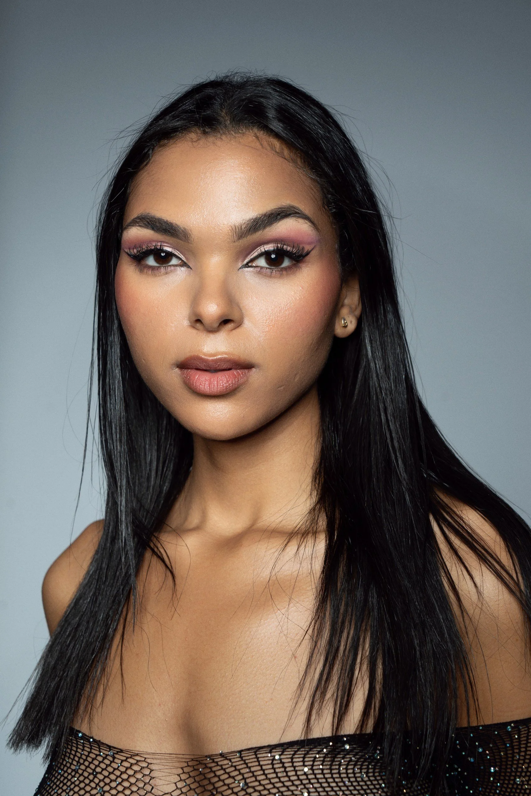 Portrait of a woman with long black hair, wearing makeup with pink eyeshadow and a mesh top, against a neutral background.