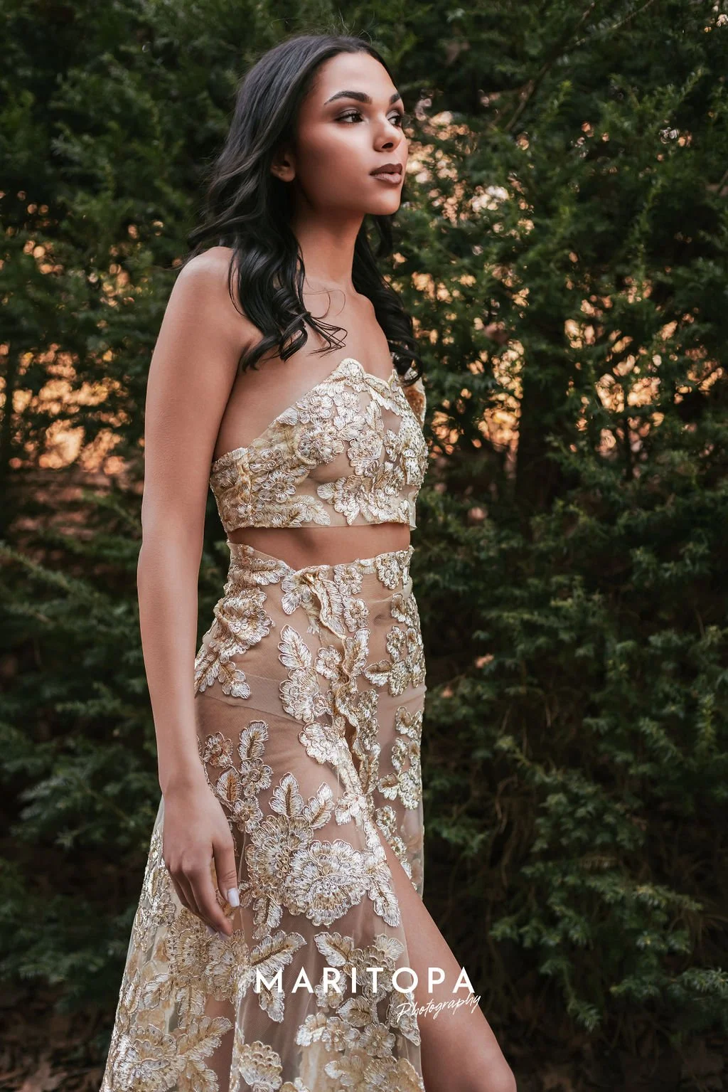 Woman in a floral embroidered two-piece formal dress standing outdoors with trees in the background.