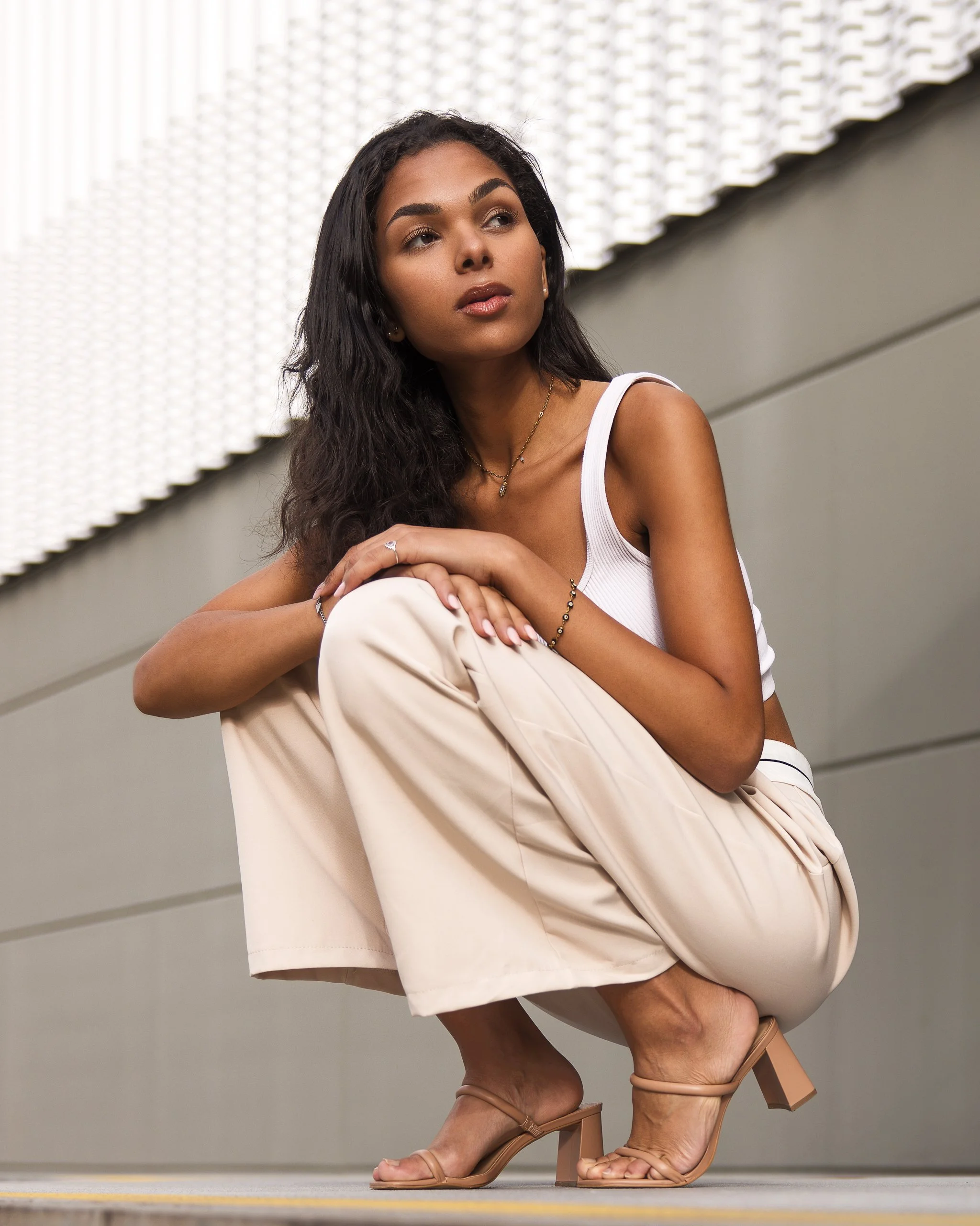 Person crouching in neutral outfit with white tank top and beige pants next to a textured wall, wearing sandals and a bracelet, looking away.