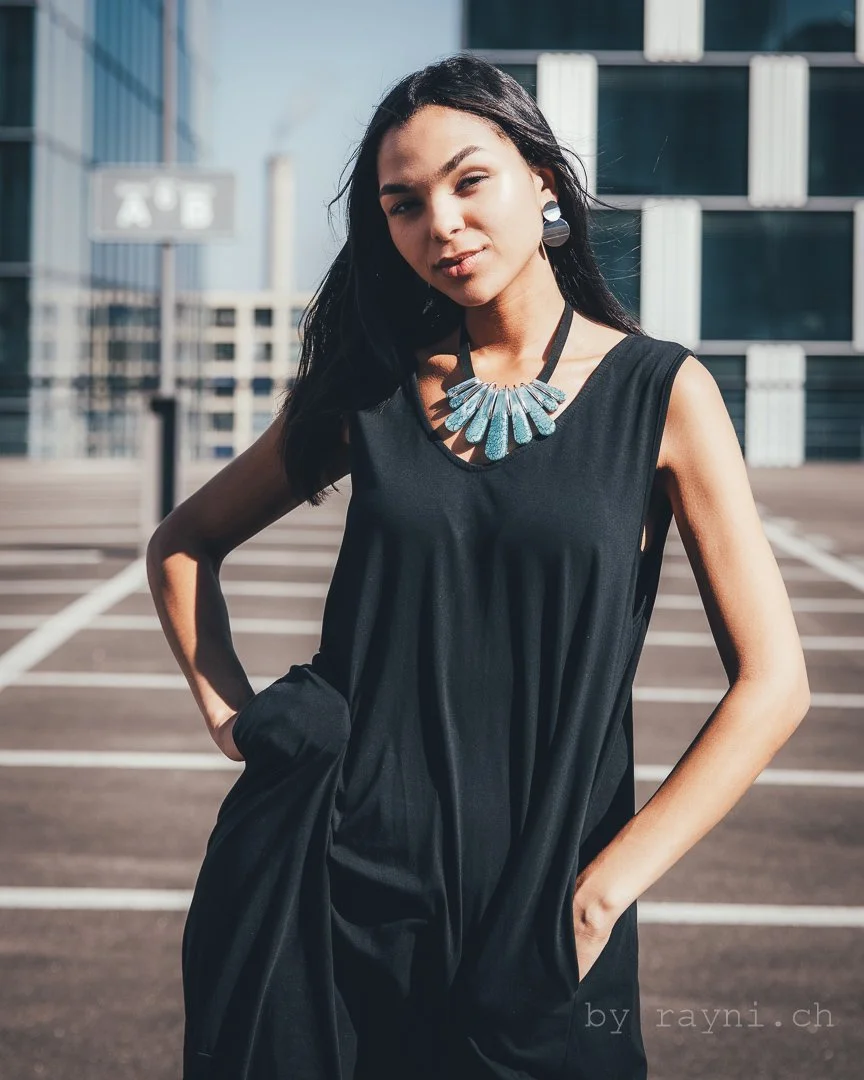 Woman wearing black dress and blue statement necklace in outdoor urban setting.