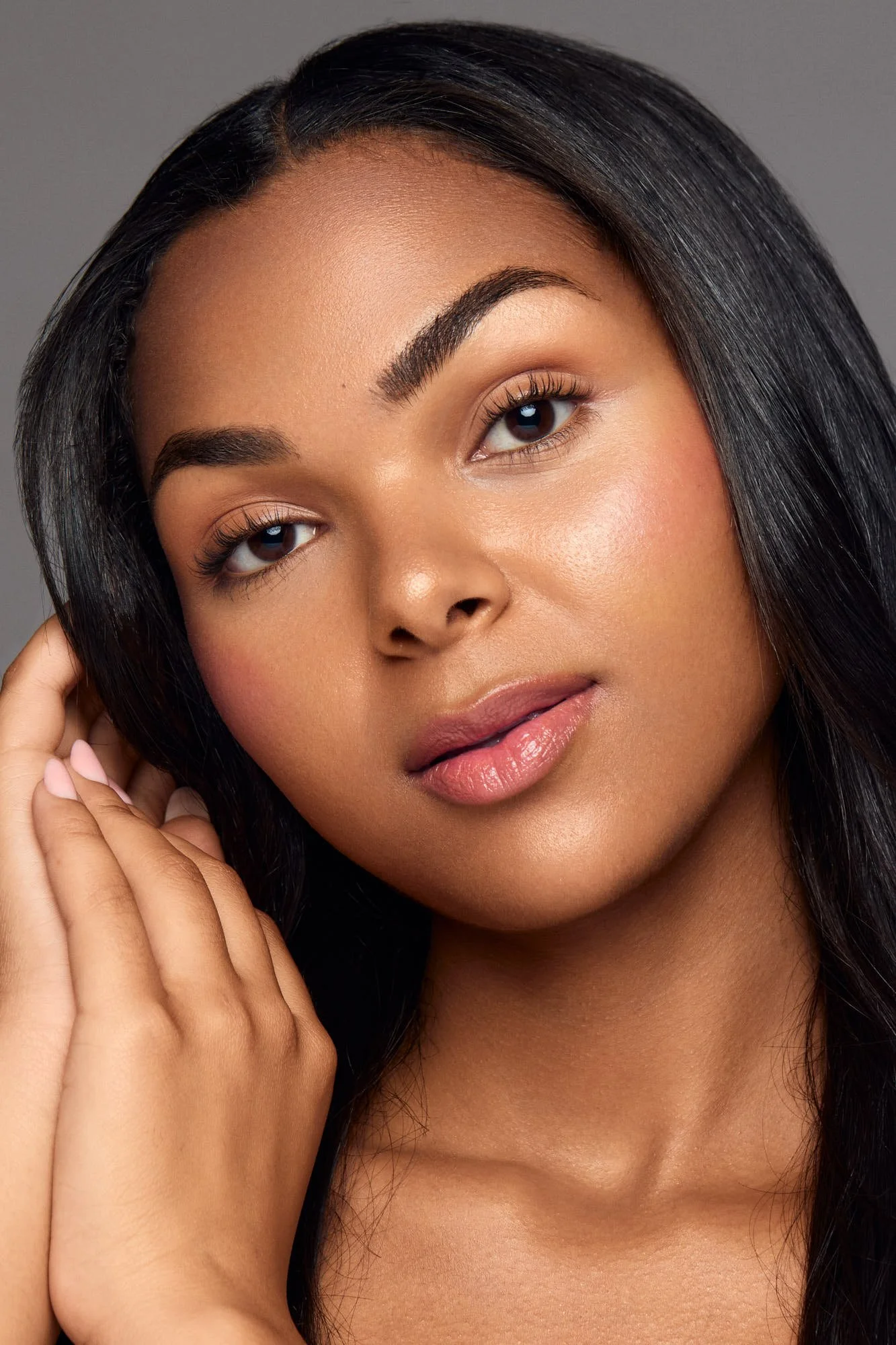 Portrait of a woman with long black hair, touching her face, and looking at the camera, showcasing natural beauty and smooth skin against a gray background.