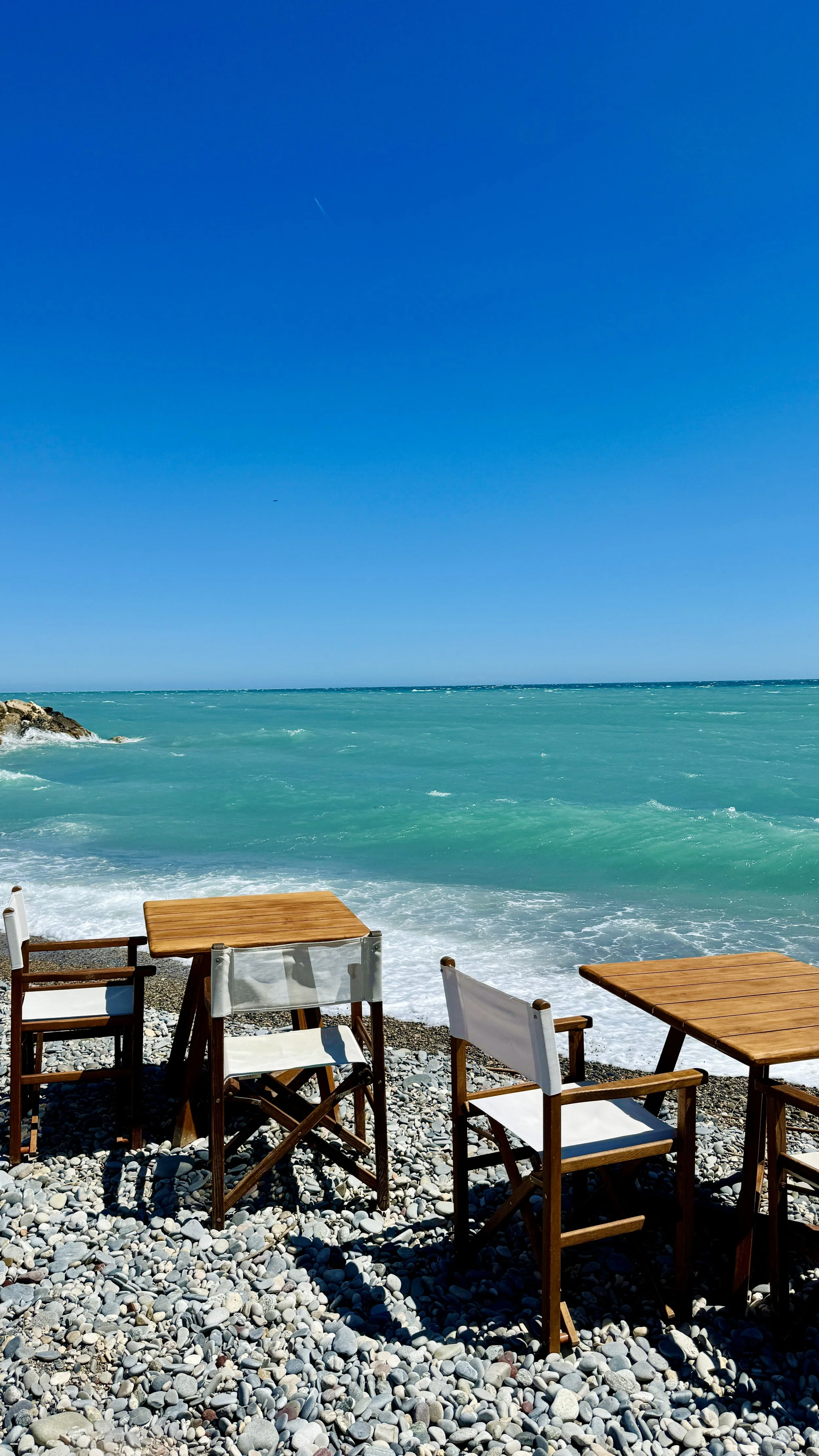 Outdoor seating area with wooden tables and chairs on a pebble beach by the sea under a clear blue sky.