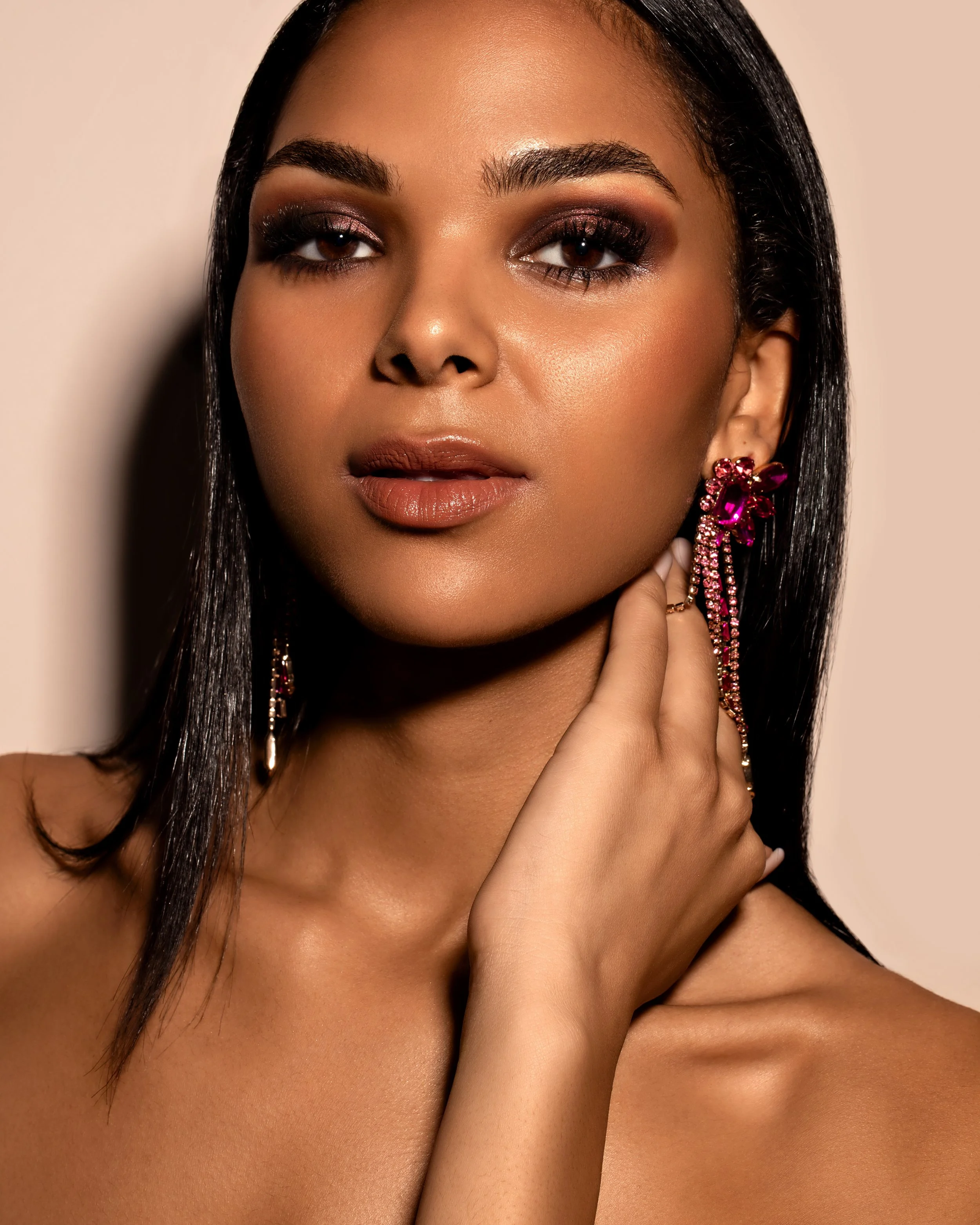 Woman with long dark hair and elegant jewelry, featuring pink dangling earrings, posing with hand near her neck, neutral background.