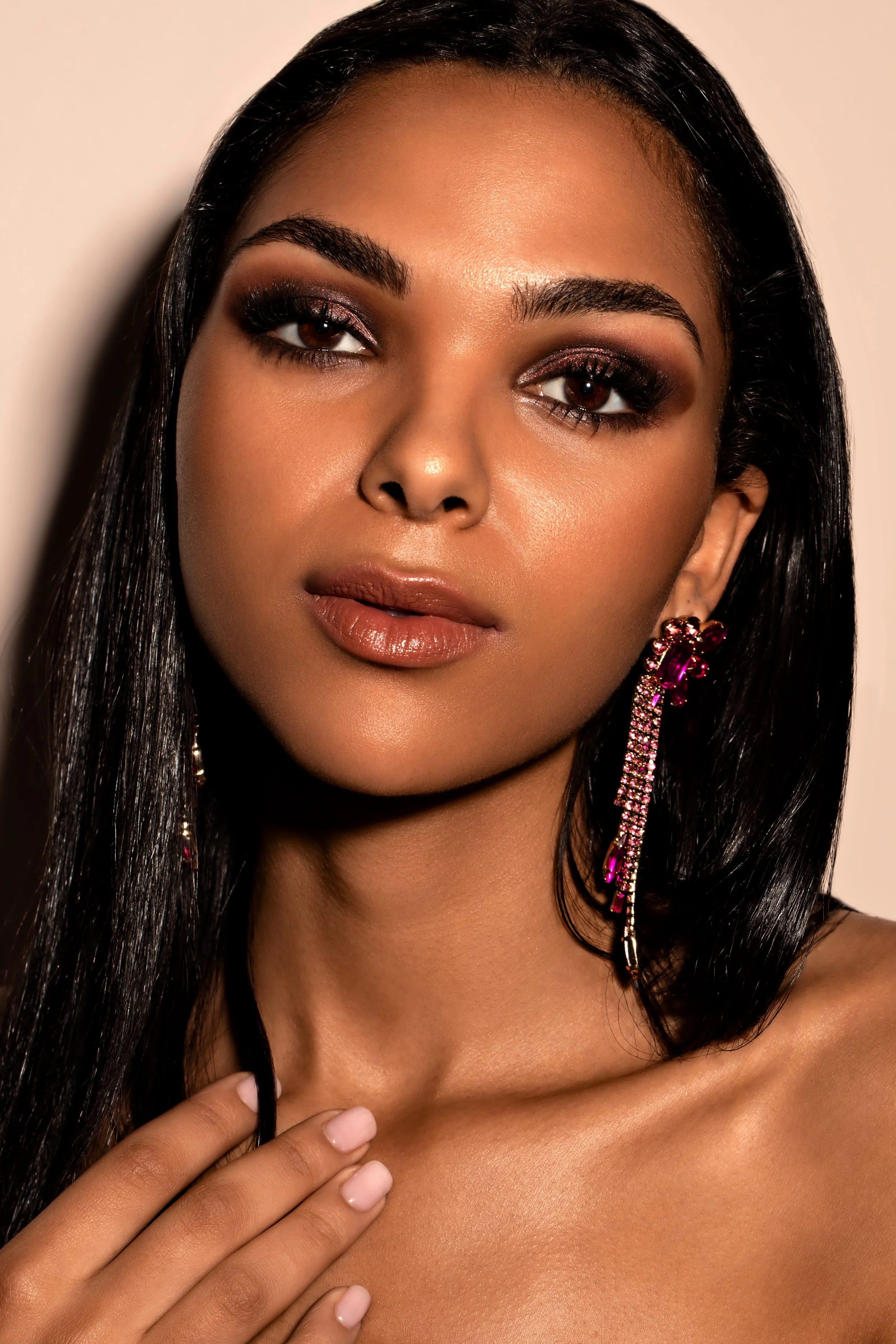 Close-up of a woman with dark hair, wearing makeup and pink earrings, touching her collarbone with manicured nails.