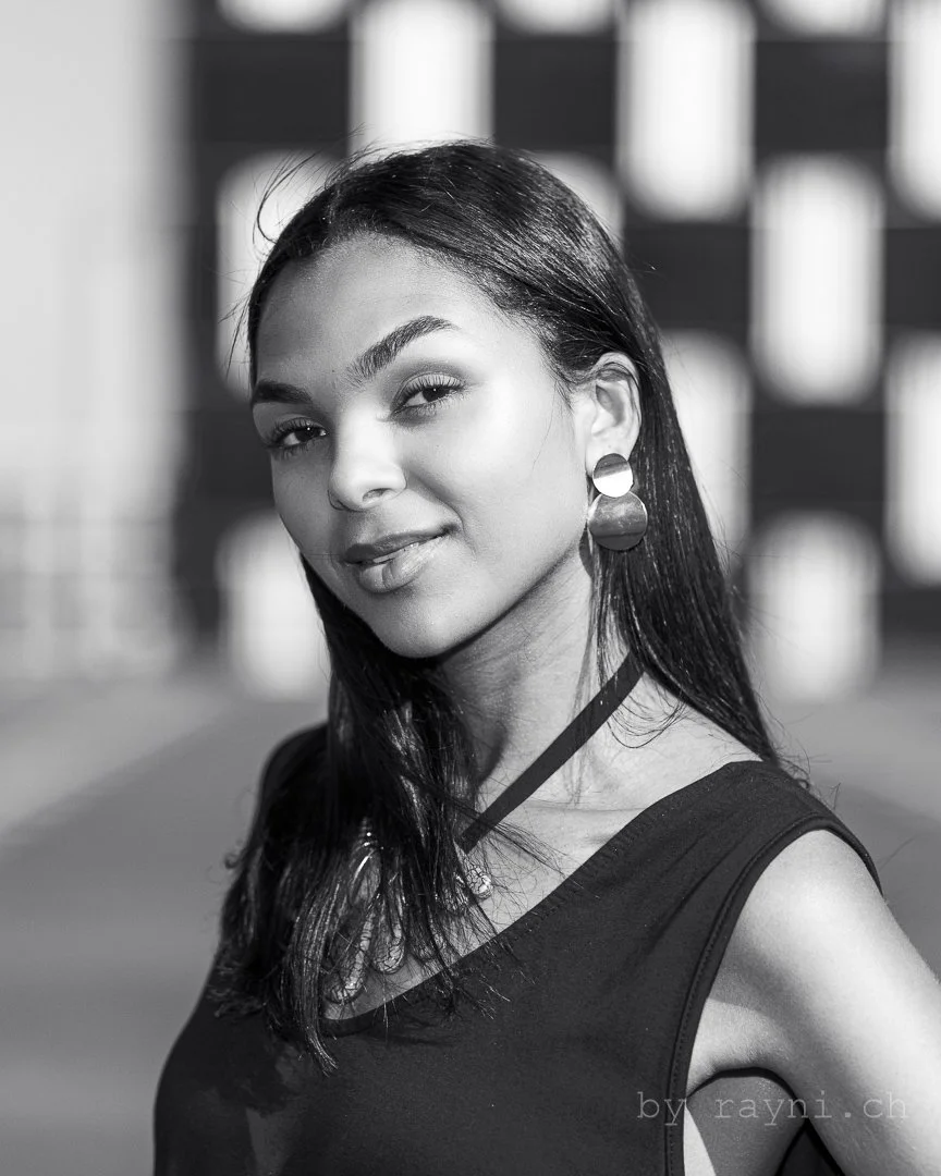 Black and white portrait of a woman with long hair wearing earrings, smiling slightly. Background is blurred with a checkered pattern.