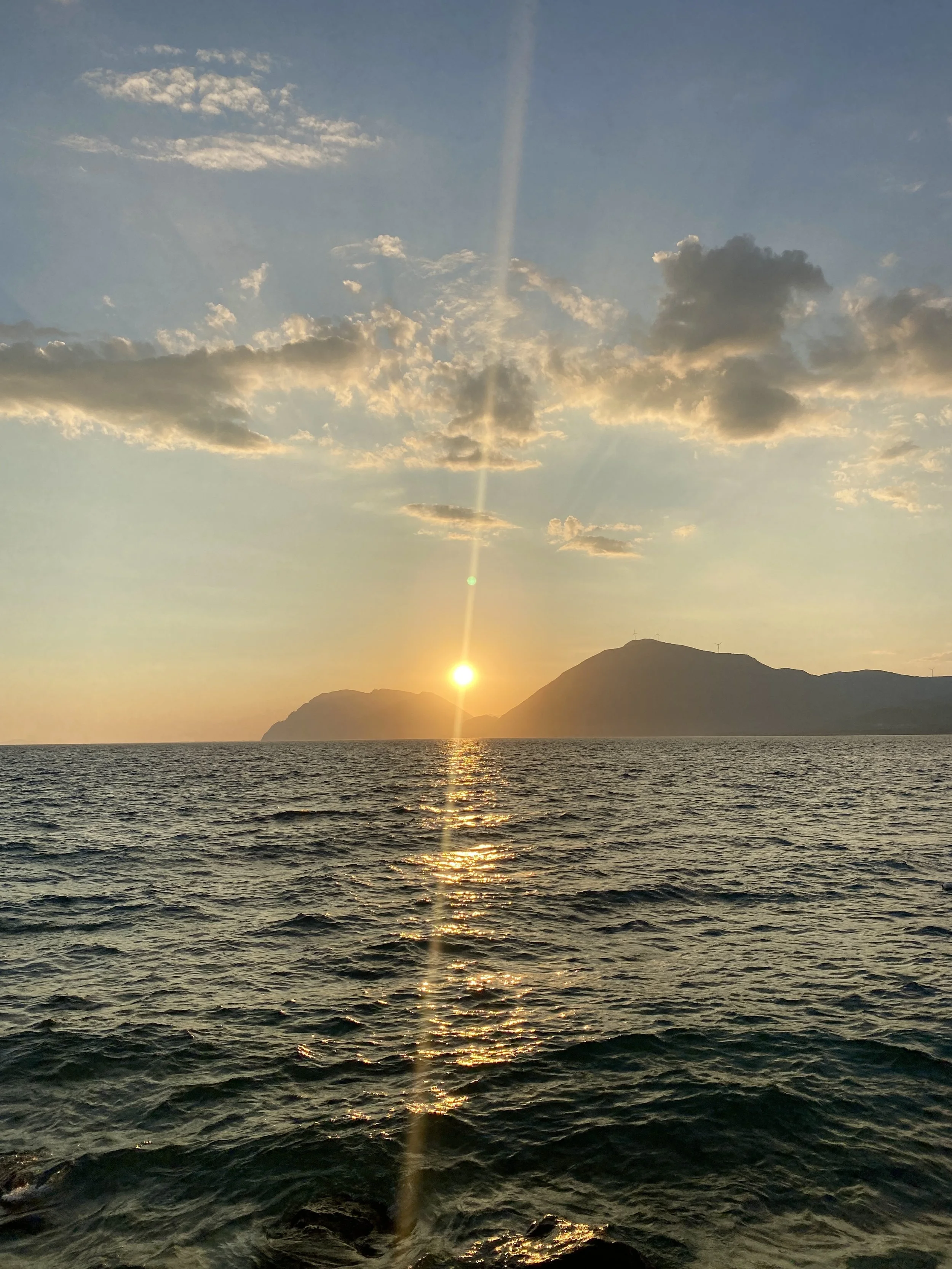 Sunset over the ocean with a mountain silhouette, scattered clouds in the sky, and sunlight reflecting on the water surface.