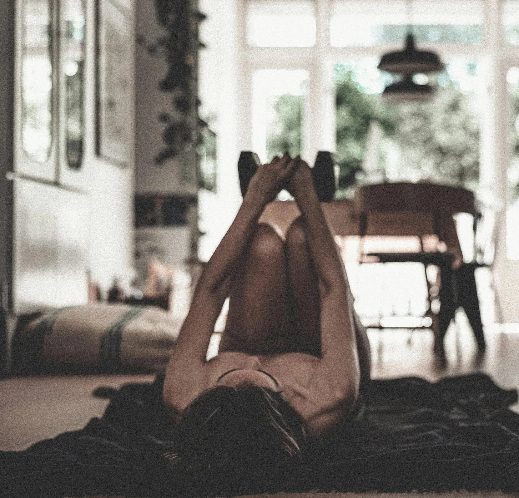 Person lying on their back on a yoga mat, holding a dumbbell with both hands, in a room with large windows and natural light.
