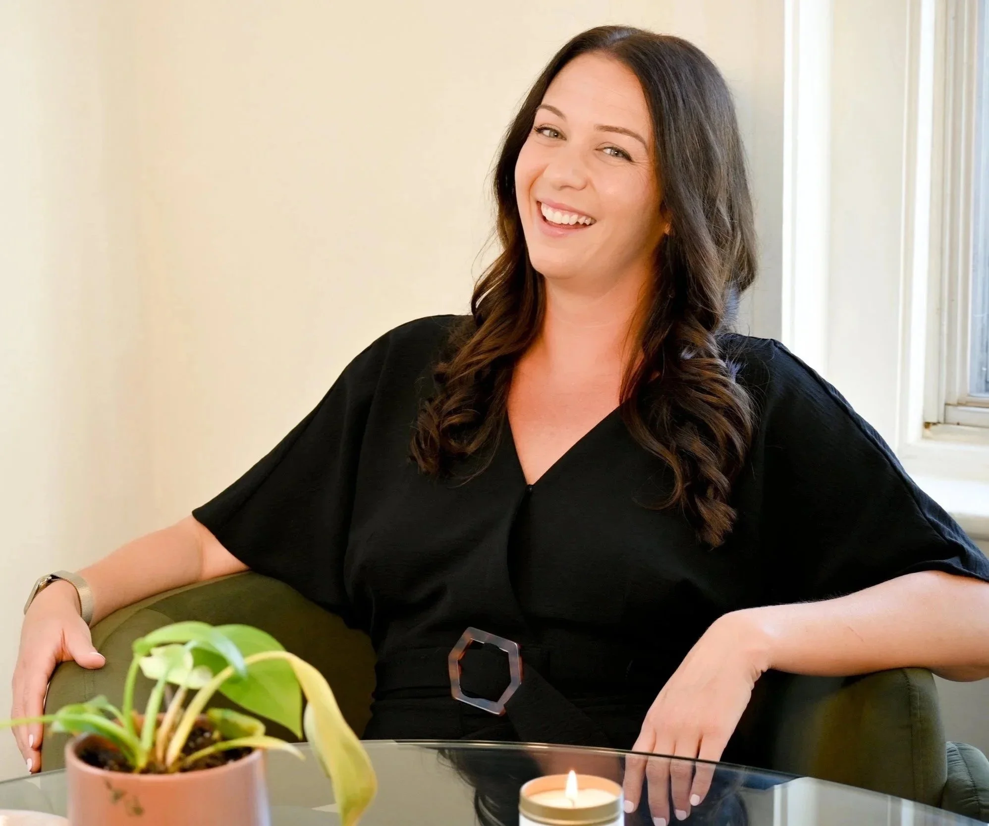 A woman with brown hair smiling while seated at a table near a window, with a potted plant and a lit candle in front of her.