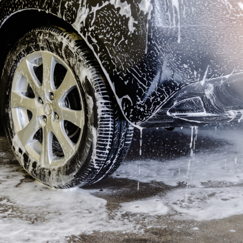 Close-up of a black car wheel being washed with soap, with soap suds and water on the tire and ground.