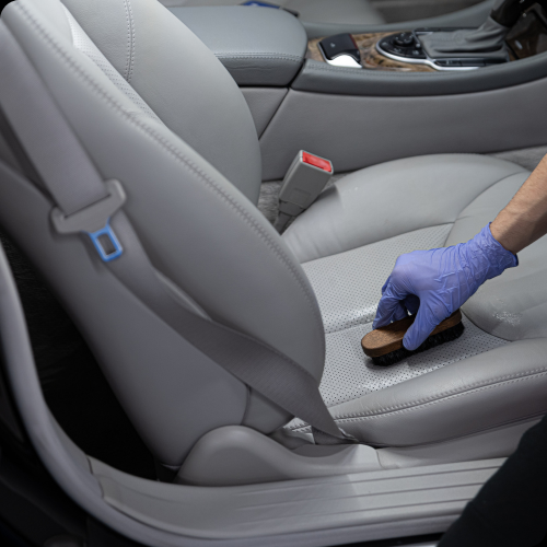 Person cleaning a car seat with a small brush while wearing a blue glove inside a modern vehicle.
