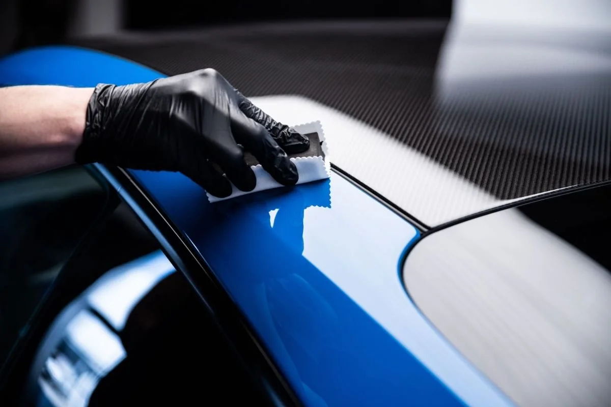 Person wearing a black glove cleaning the surface of a blue car with a white cloth.