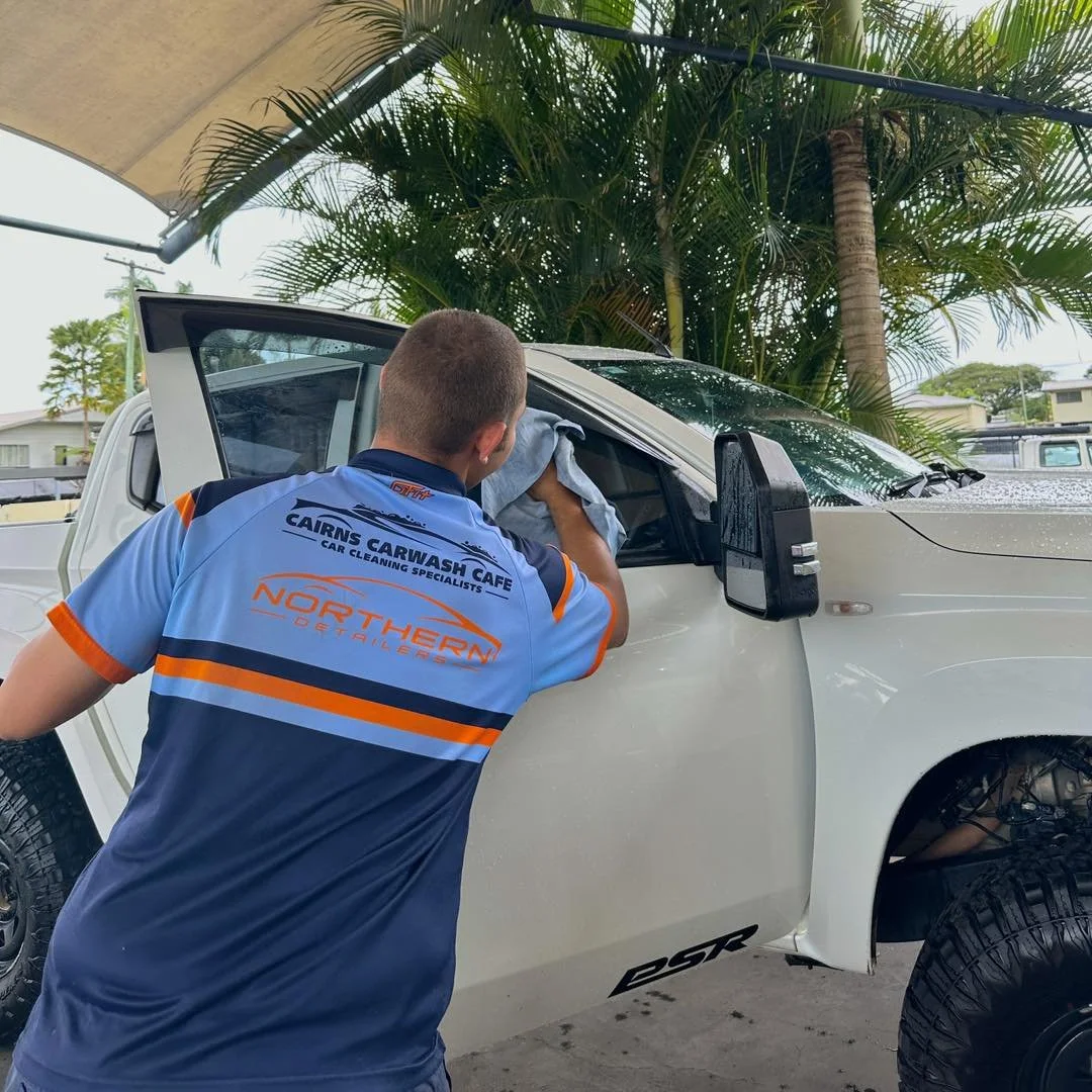 A car wash worker cleaning a white pickup truck under a canopy with palm trees in the background.