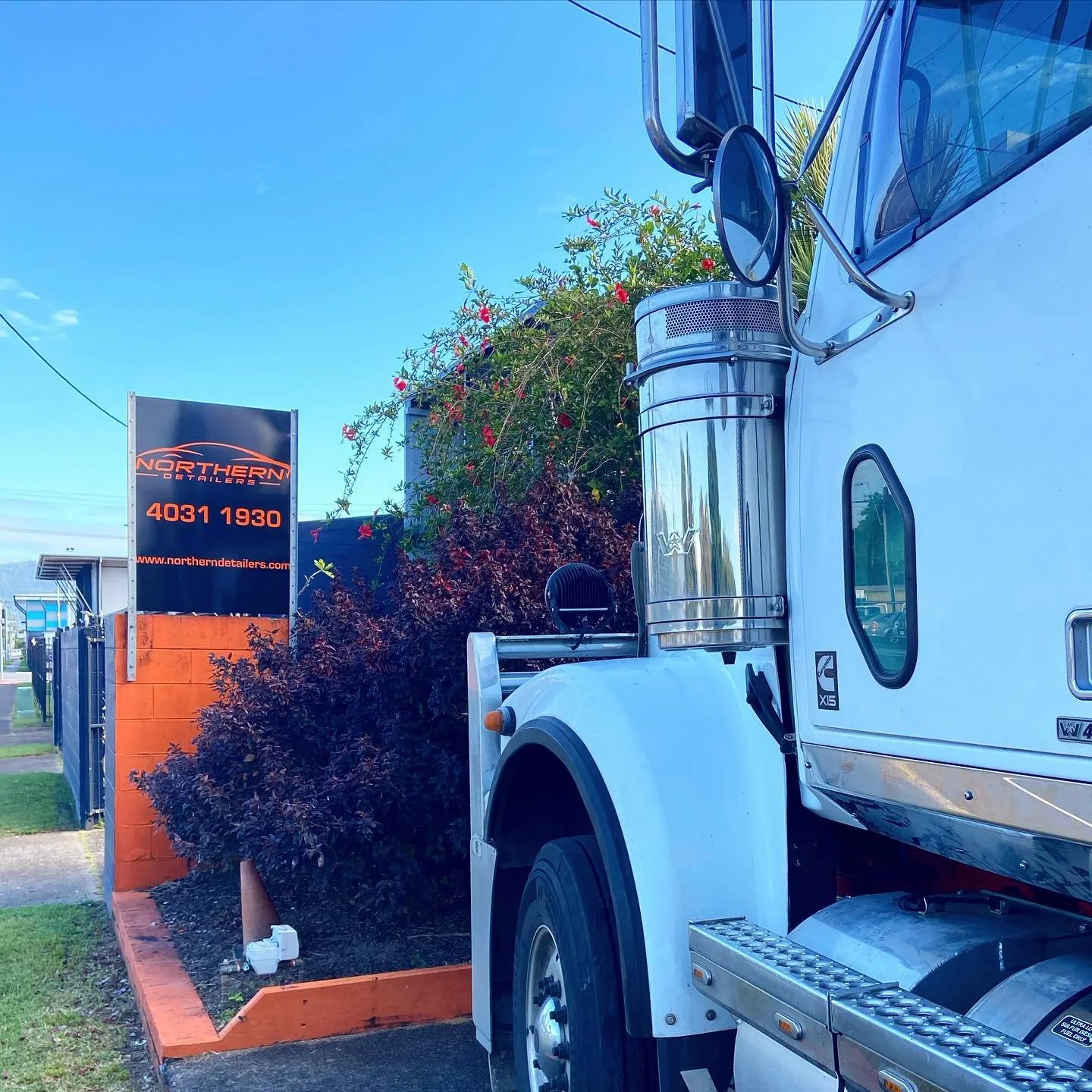 Part of a large white truck beside a black sign with orange and white lettering for Northern Detailing and a phone number, next to a well-maintained garden bed with dark purple shrubbery and red flowers, under a clear blue sky.
