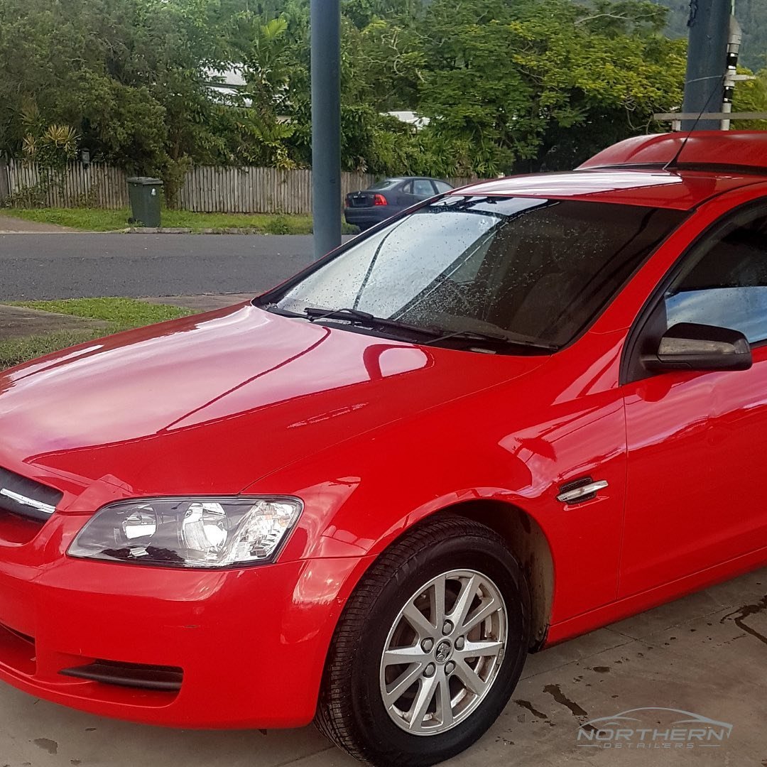 A red car is parked on a driveway with rain droplets on the windshield. The background includes trees, a street, and a wooden fence.