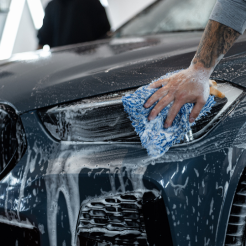 Person washing a dark-colored car with soap and a blue sponge.